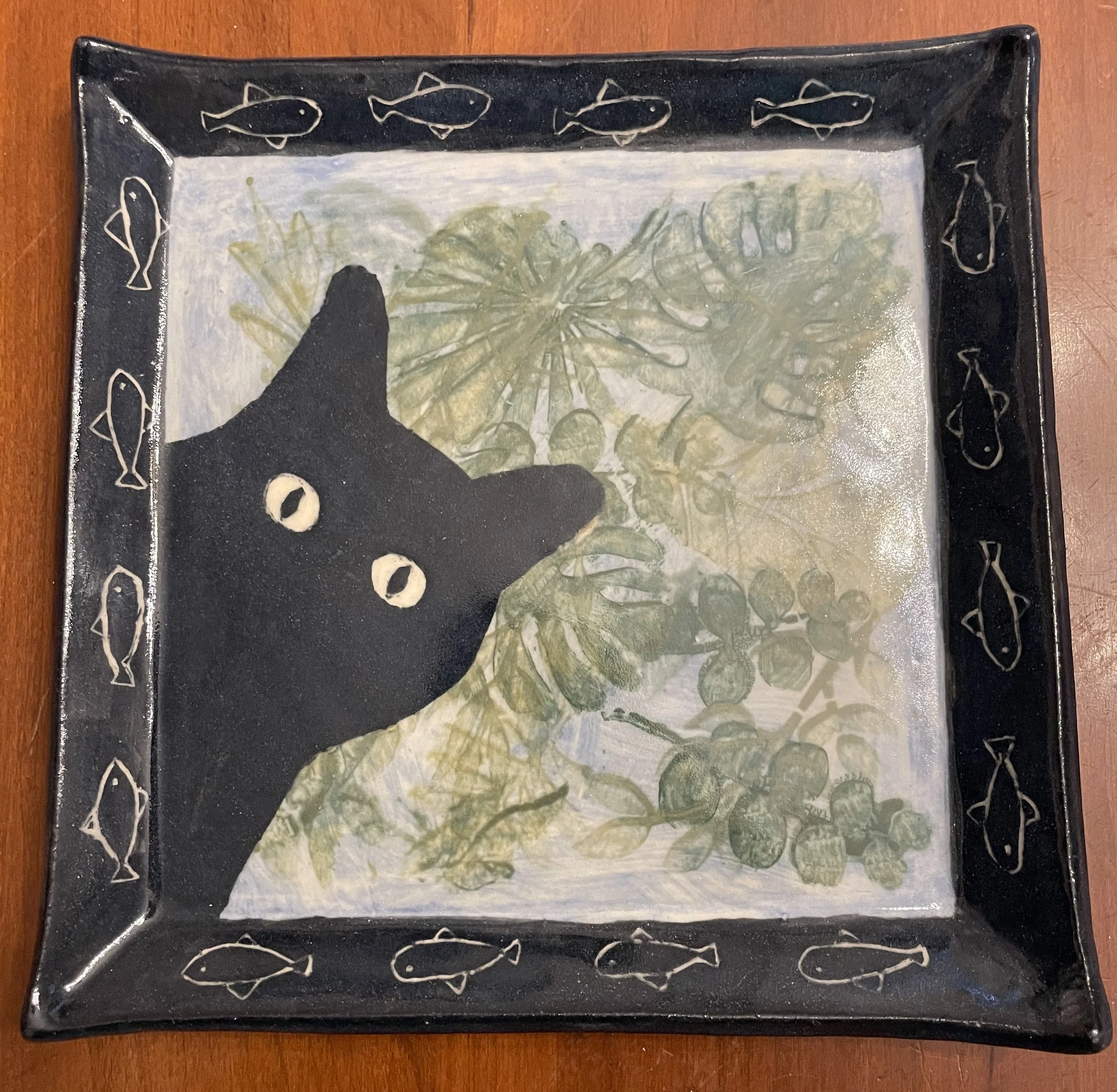 A square black tray with a fish pattern on the border, showing a black cat silhouette with wide eyes in the foreground, and a background of green leafy plants and berries.