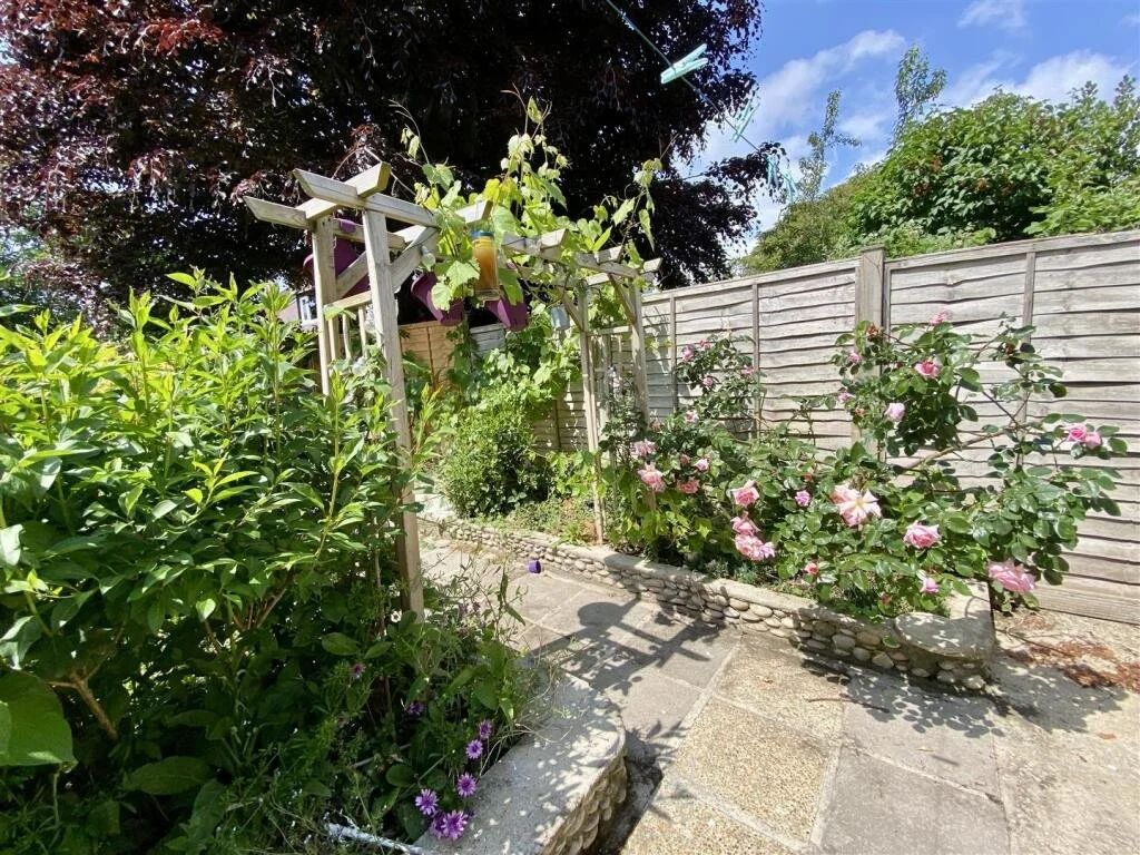 A small backyard garden with a wooden fence, blooming pink and purple flowers, green shrubbery, and a potted plant hanging from a pergola.