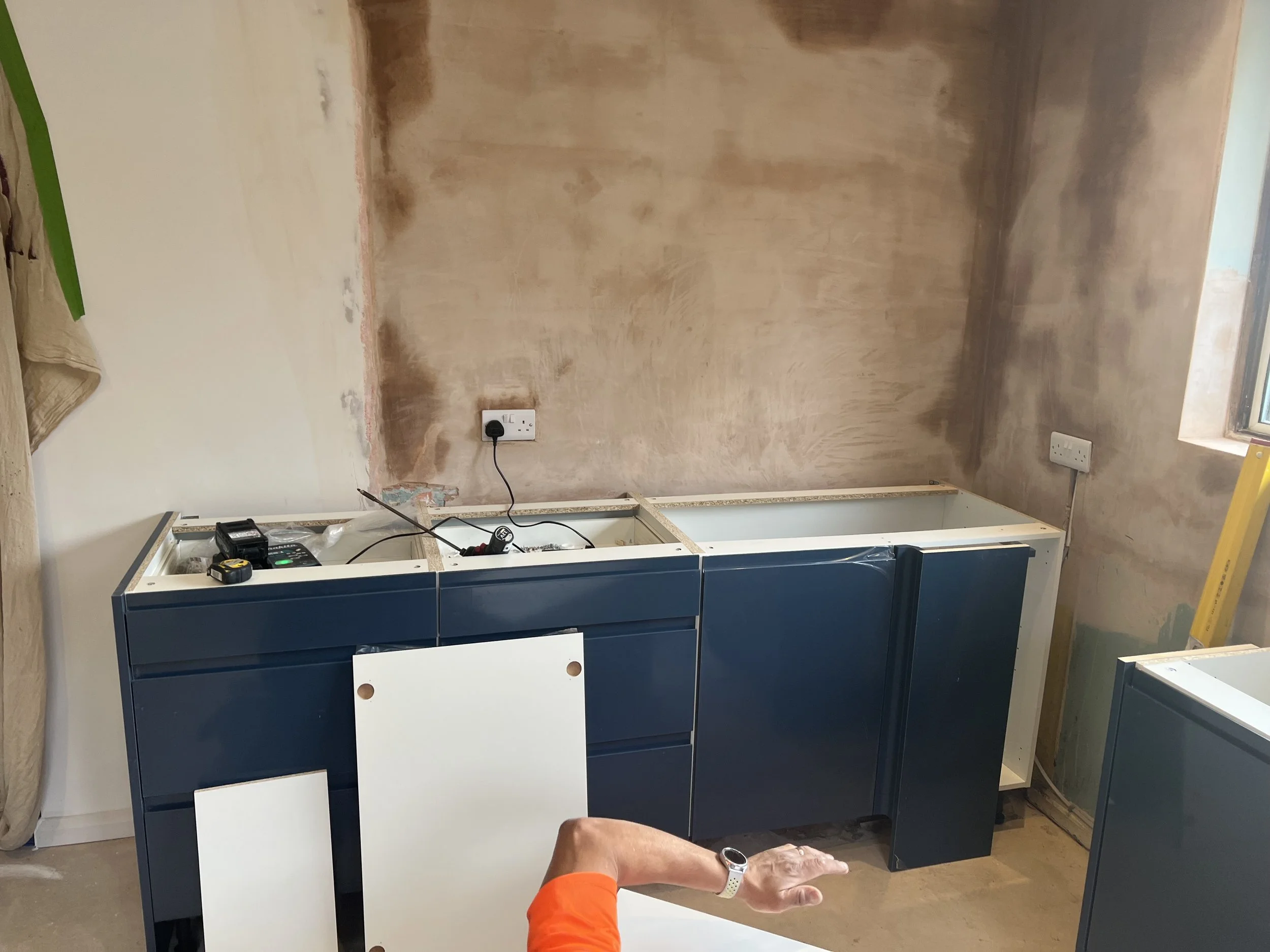 Kitchen renovation in progress with blue cabinets installed, unfinished wall with patches and electrical outlets, construction tools on the counter, and a person’s hand pointing at the cabinets.