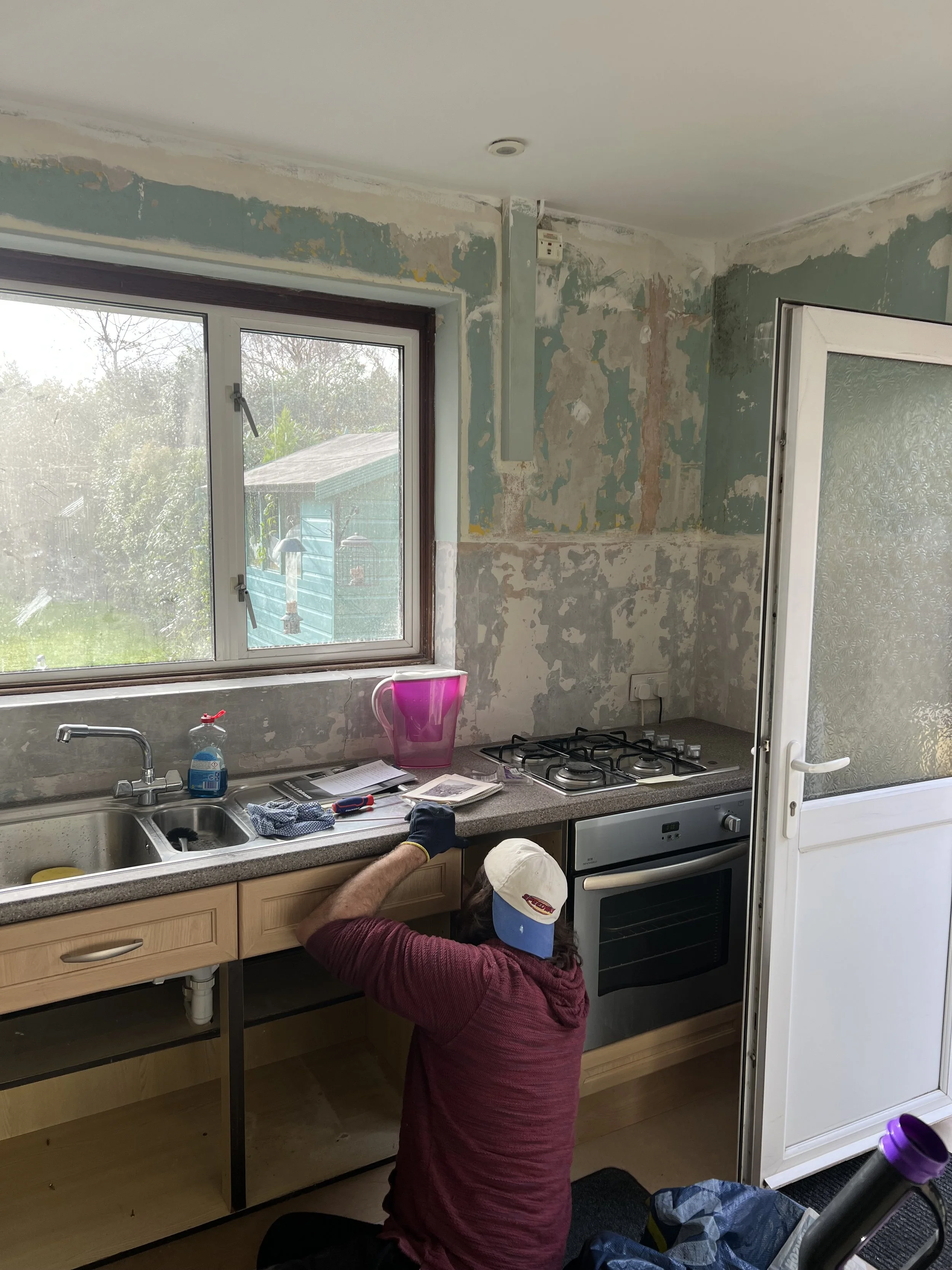 A person working on a kitchen renovation, installing or repairing the cabinetry under a window with peeling paint on the walls. The person is wearing a baseball cap and gloves, and is crouched down next to a countertop with various tools and material