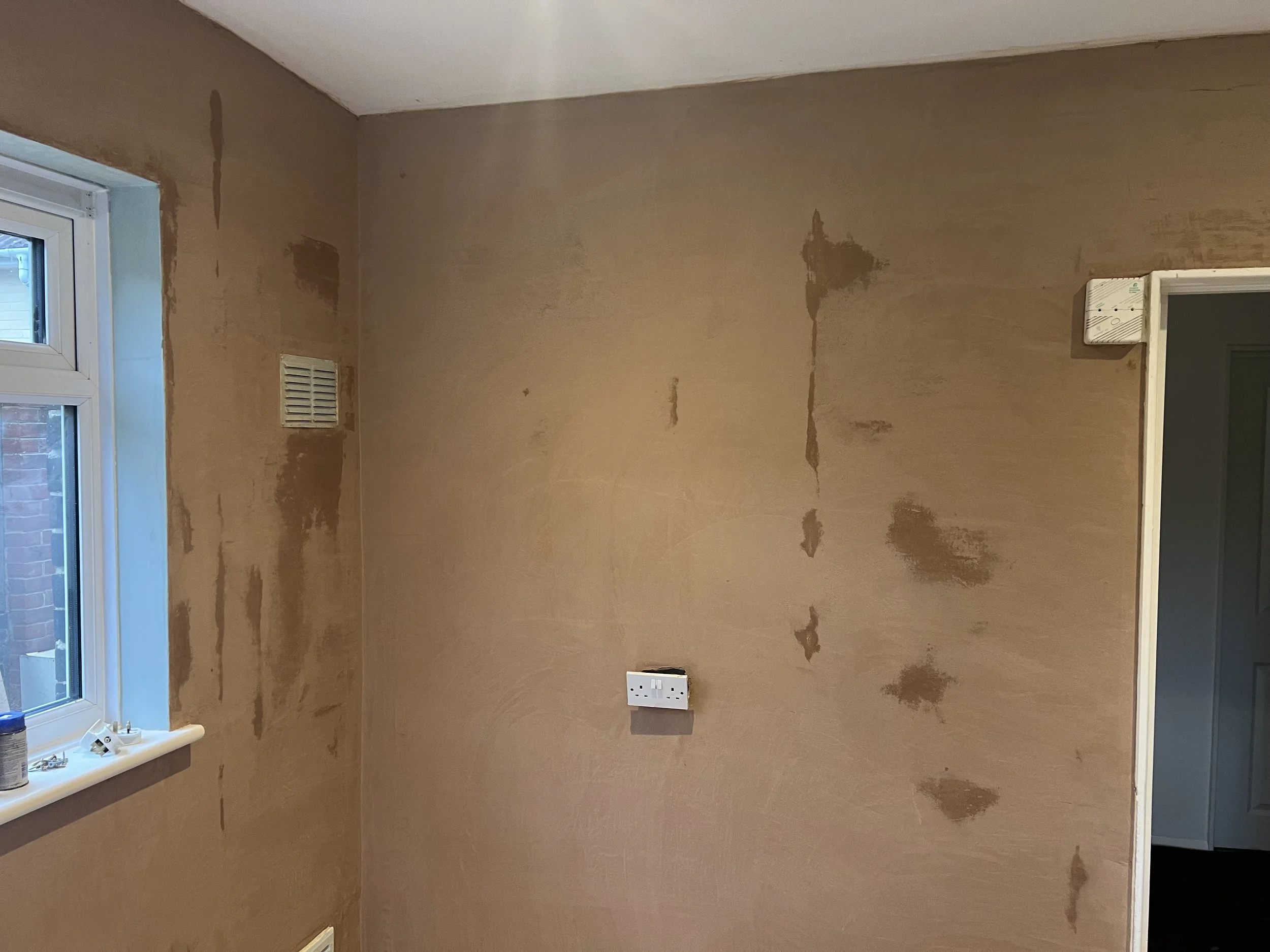 A room with stained and unpainted beige walls, a window on the left, an electrical outlet on the wall, and an open doorway on the right leading to another room.