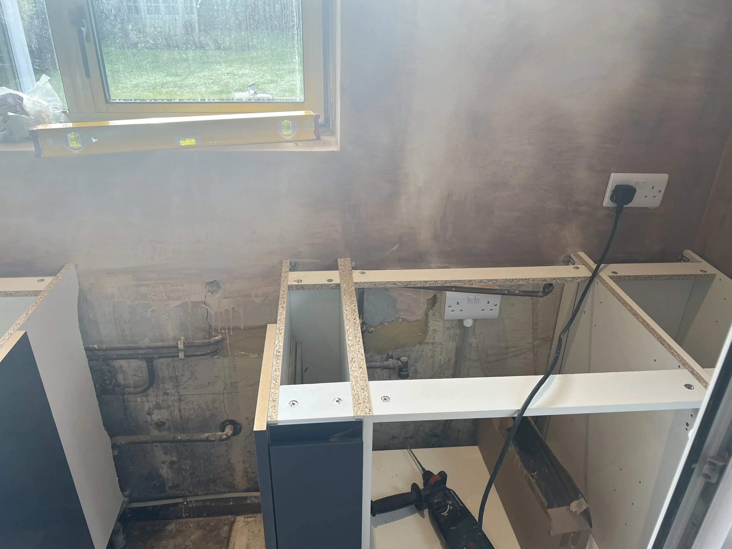 Kitchen undergoing renovation, with empty white cabinets, partially installed, a black power drill on the floor, a yellow level on the window sill, and electrical outlets on the wall.
