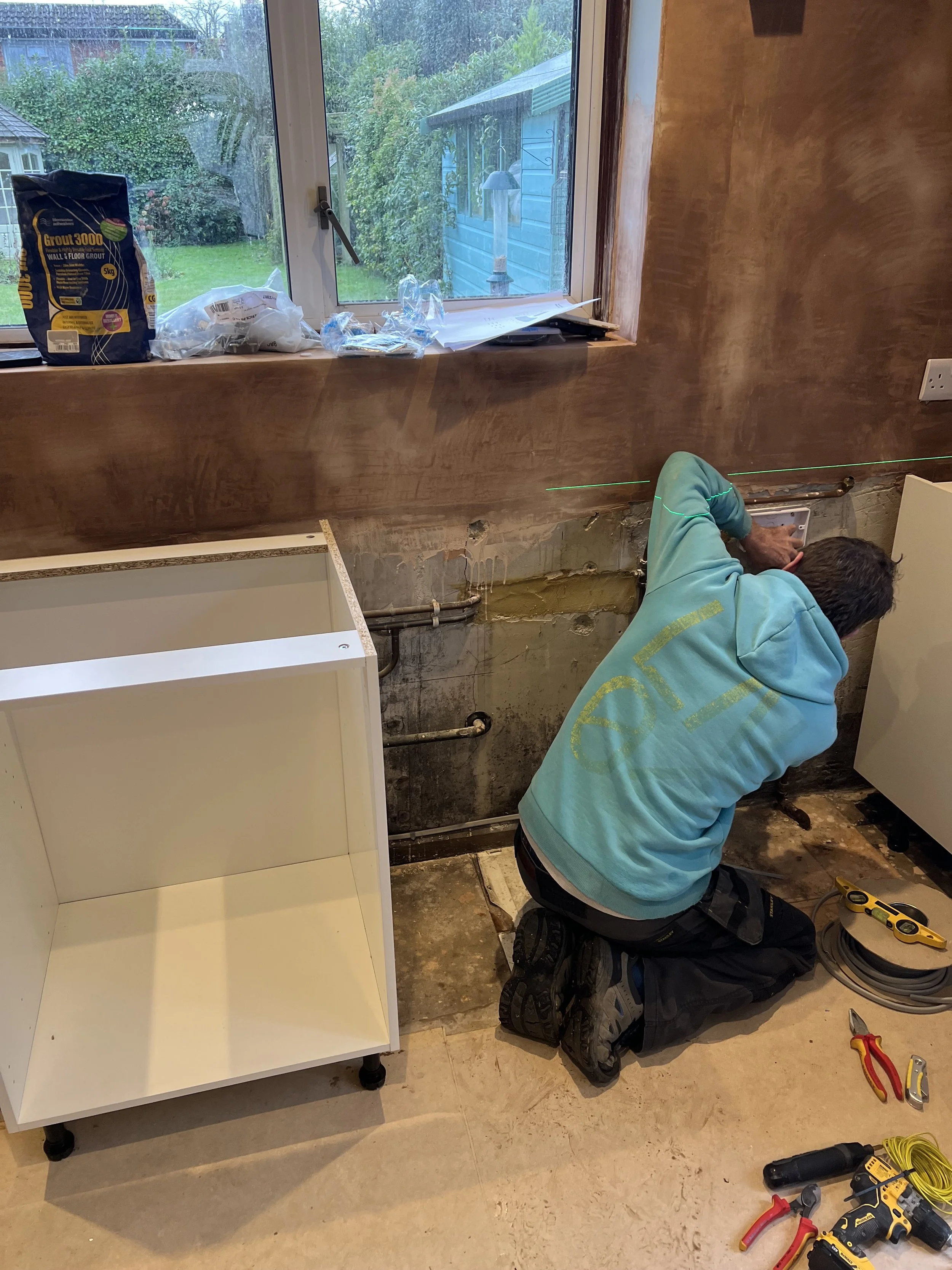 A person kneeling on the floor working on plumbing or electrical wiring in a kitchen with an unfinished wall and a window showing a backyard.