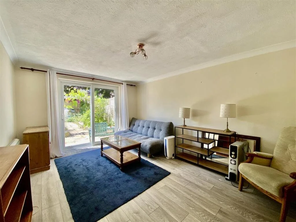 Living room with sliding glass door leading outside, featuring a gray couch, a wooden coffee table on a navy blue rug, a wooden side table, a TV stand with two lamps, a beige armchair, and wooden flooring.