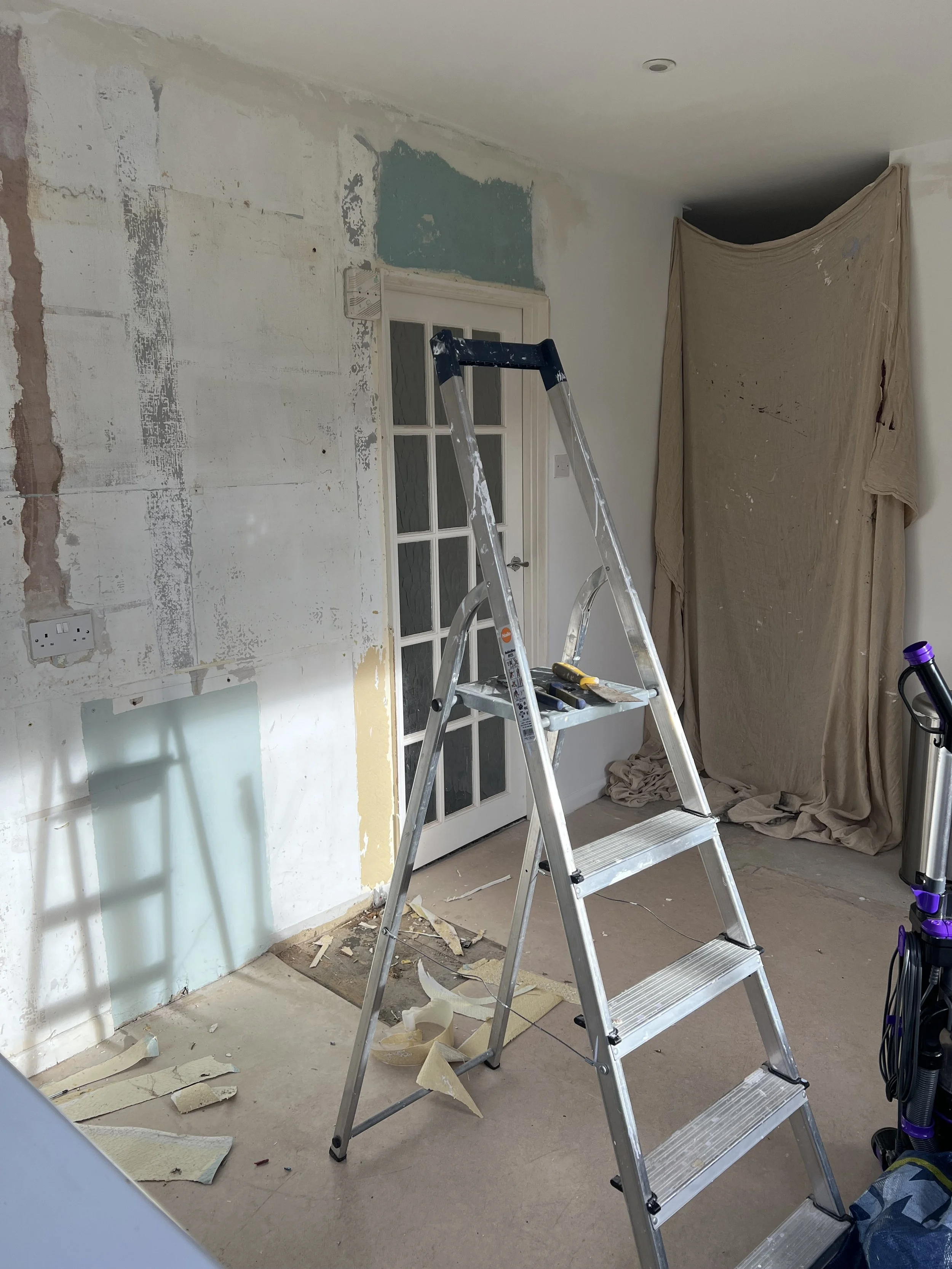 Room under renovation with a ladder, torn wallpaper, and plastic sheeting covering part of the wall.