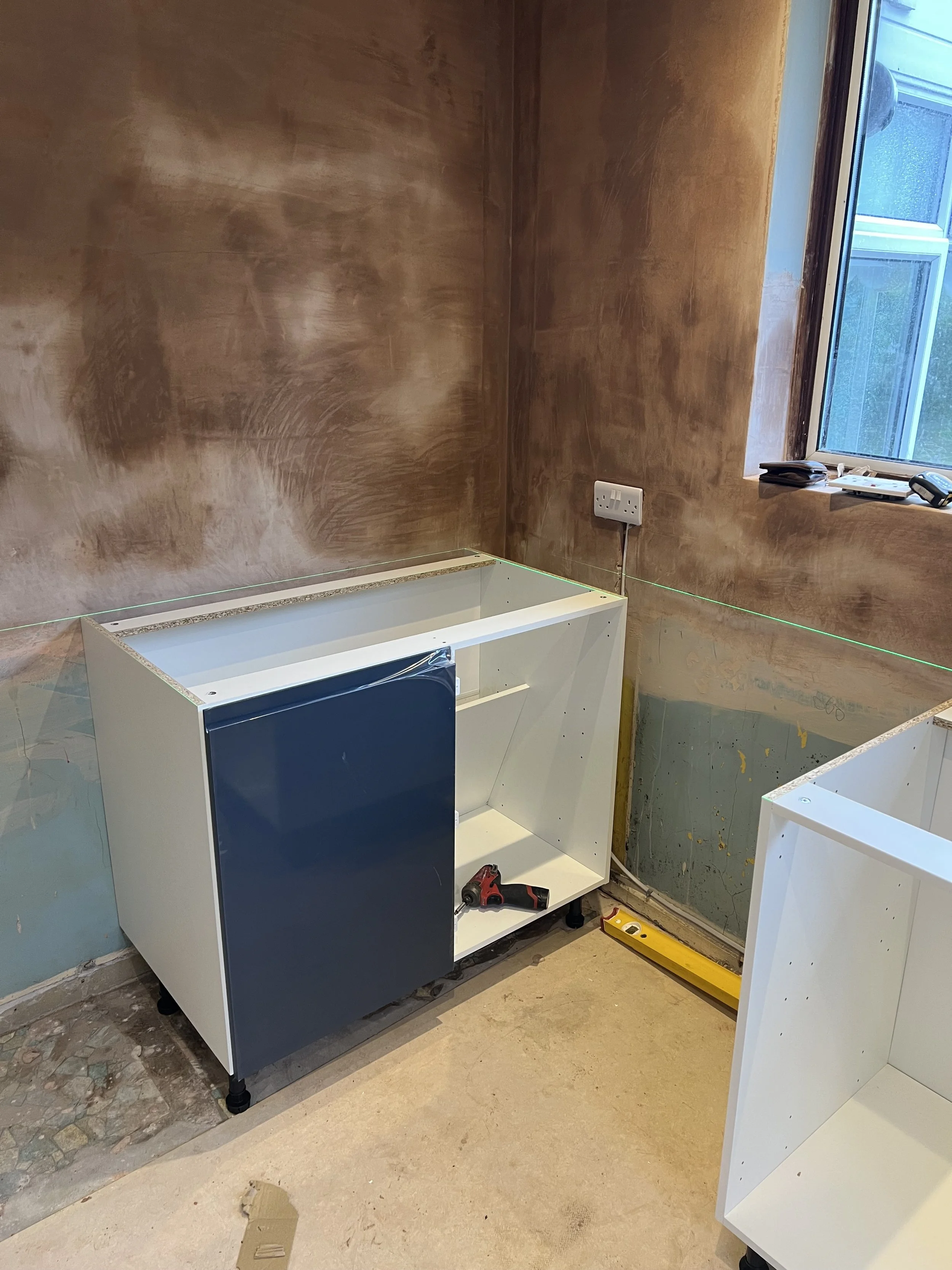 Partial kitchen or cabinetry installation with a white cabinet that has a blue door, tools, and an unfinished floor, in a room with brown walls and a window.