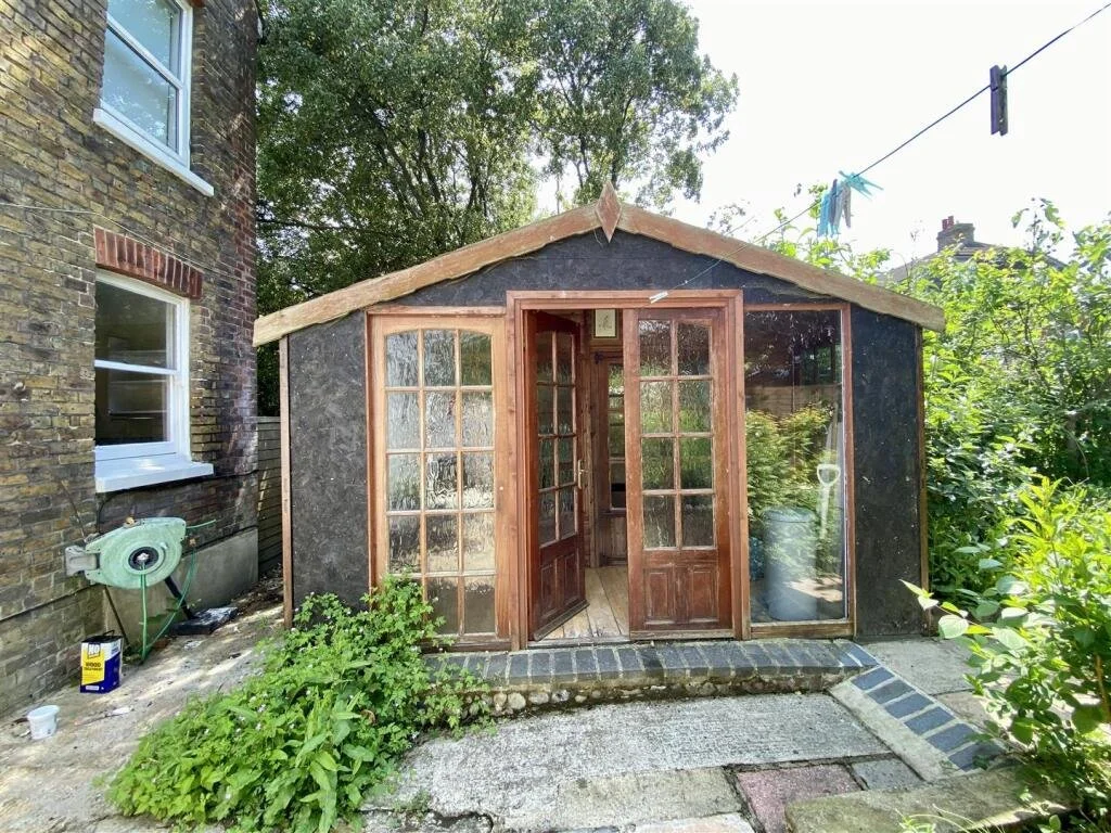 Small wooden greenhouse with open glass-paneled doors and large windows, surrounded by greenery and plants, adjacent to a brick building.