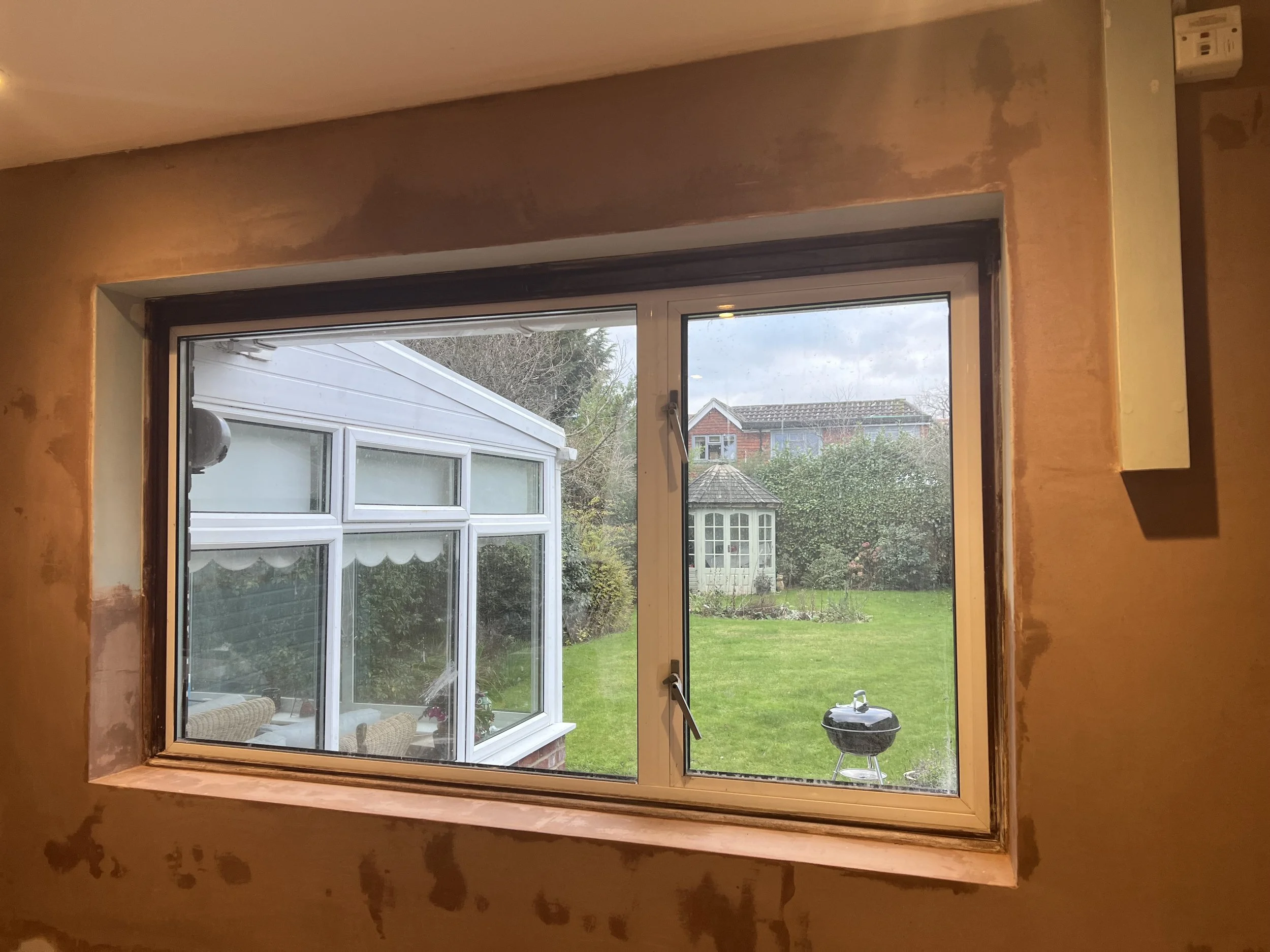 View through a window showing a green backyard with a grassy lawn, a small white garden shed, a barbecue grill, and neighboring houses in the distance.