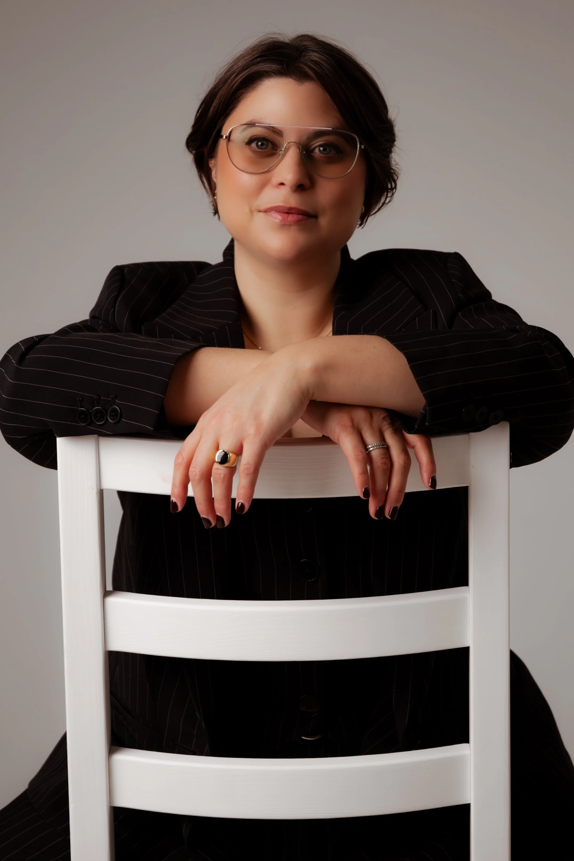 A woman with short dark hair wearing glasses and a black pinstripe suit, sitting behind a white chair, resting her arms on the chair back, looking at the camera.