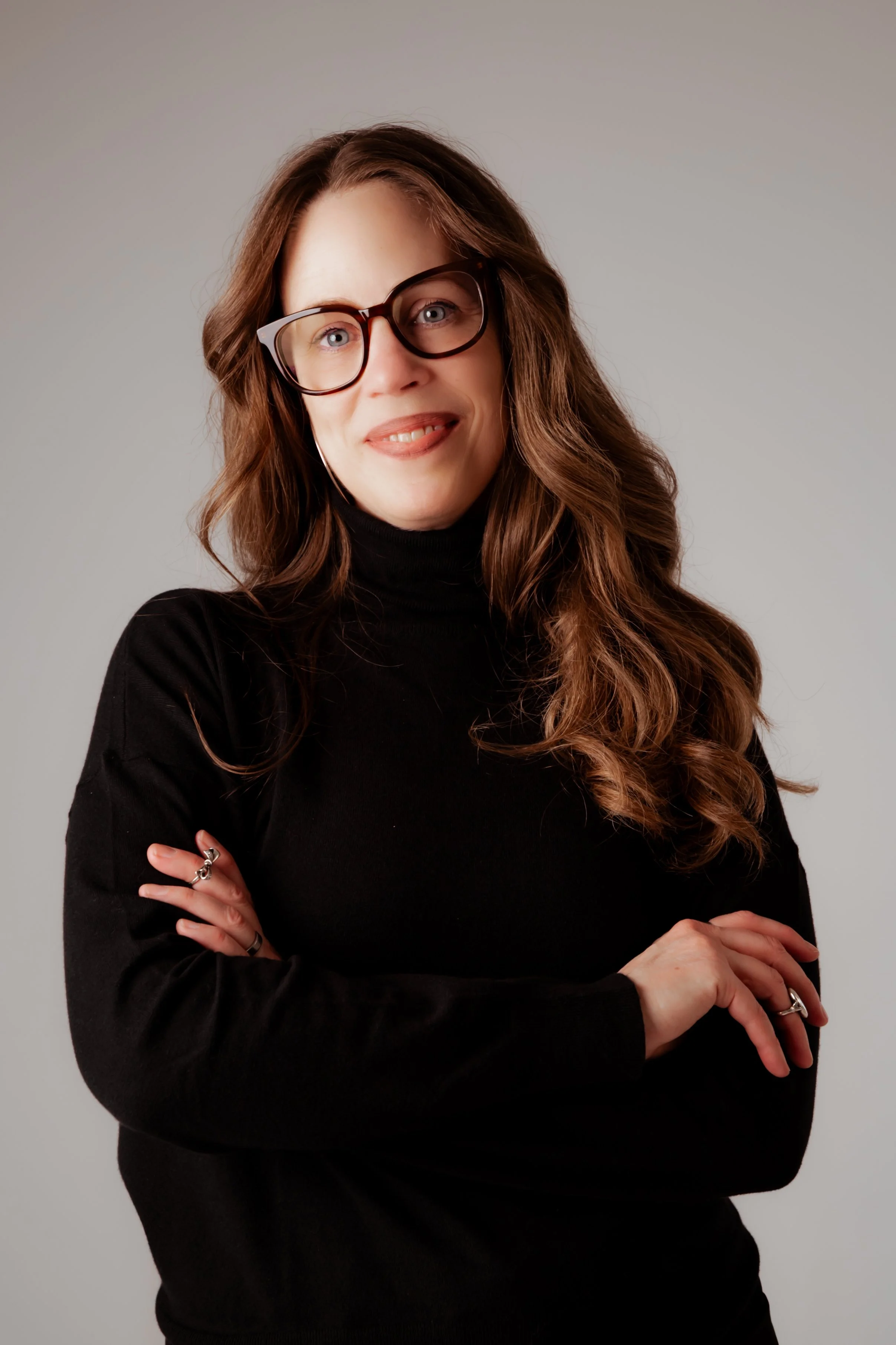 A woman with long wavy brown hair, wearing glasses and a black turtleneck, crosses her arms and smiles at the camera against a plain background.