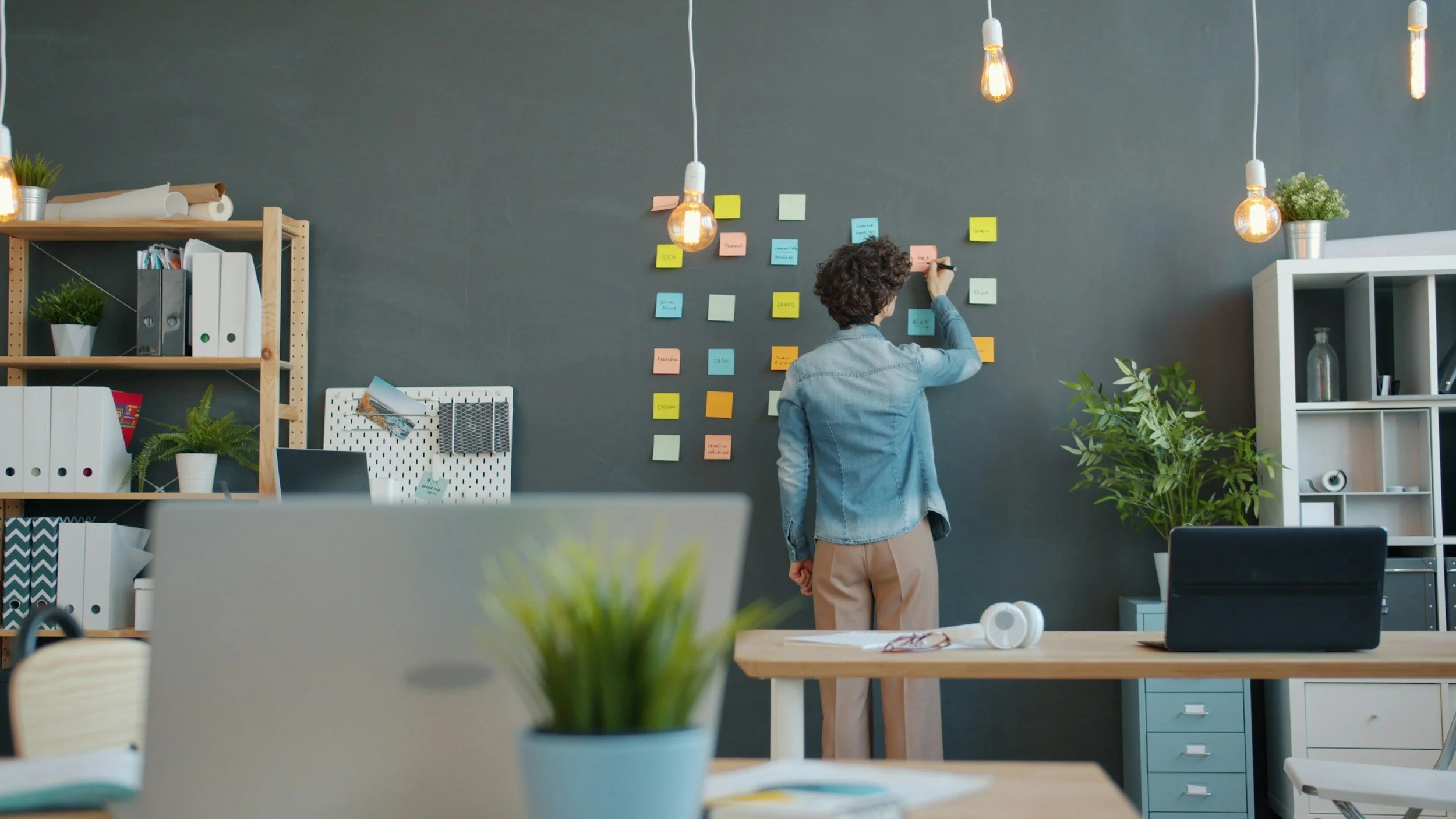 Person standing and writing on sticky notes on a dark gray wall in an office or workspace, with plants, shelves, and office supplies nearby.