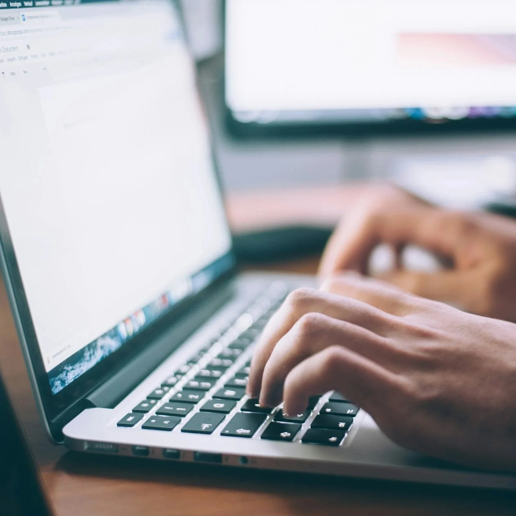 Close-up of hands typing on a laptop keyboard with a blurred computer screen in the background.