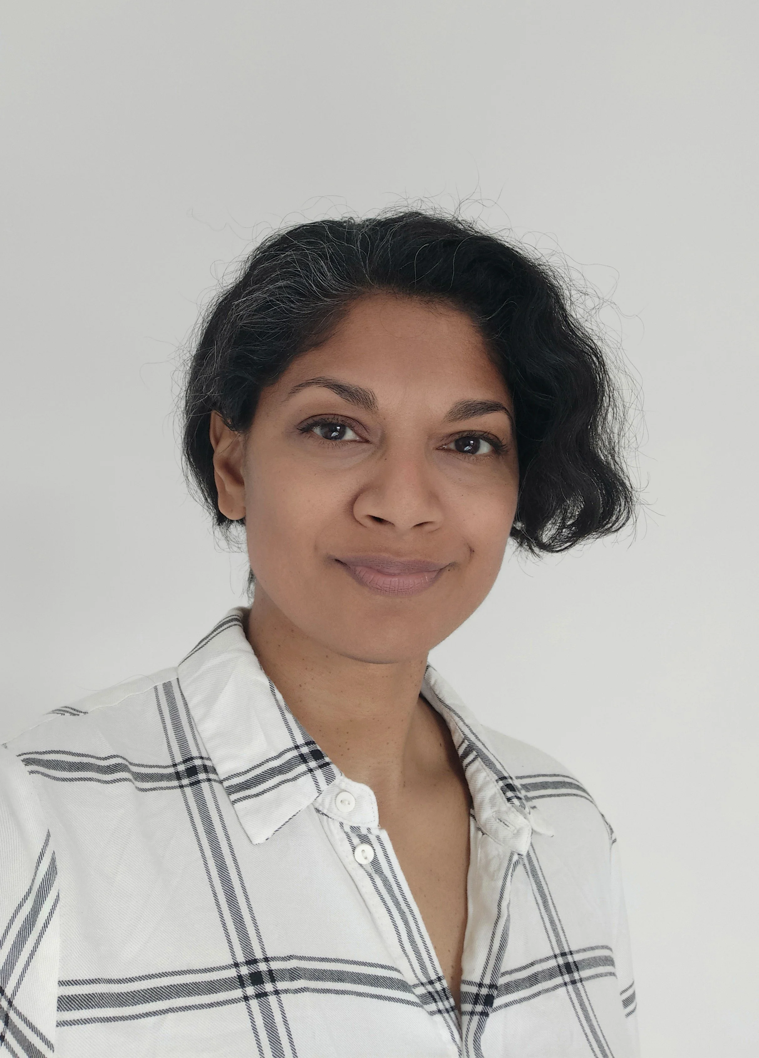 Picture of the founder, Sharon Addenbrooke with short, wavy black hair wearing a white shirt with black stripes, standing against a plain white wall.