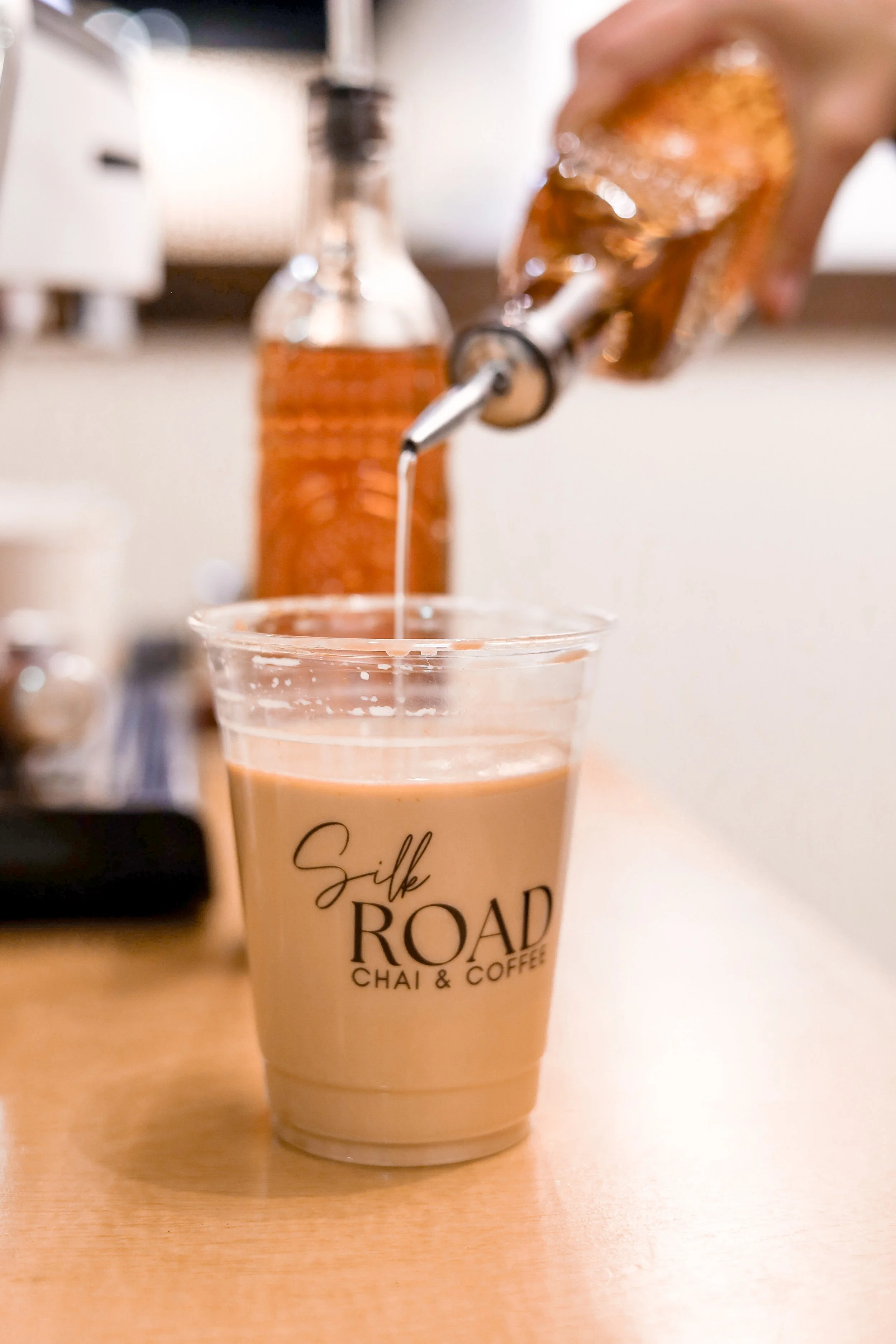 A plastic cup of chai tea with milk, on a wooden table, labeled 'Silk Road Chai & Coffee', with honey or syrup being poured into it from a squeeze bottle.