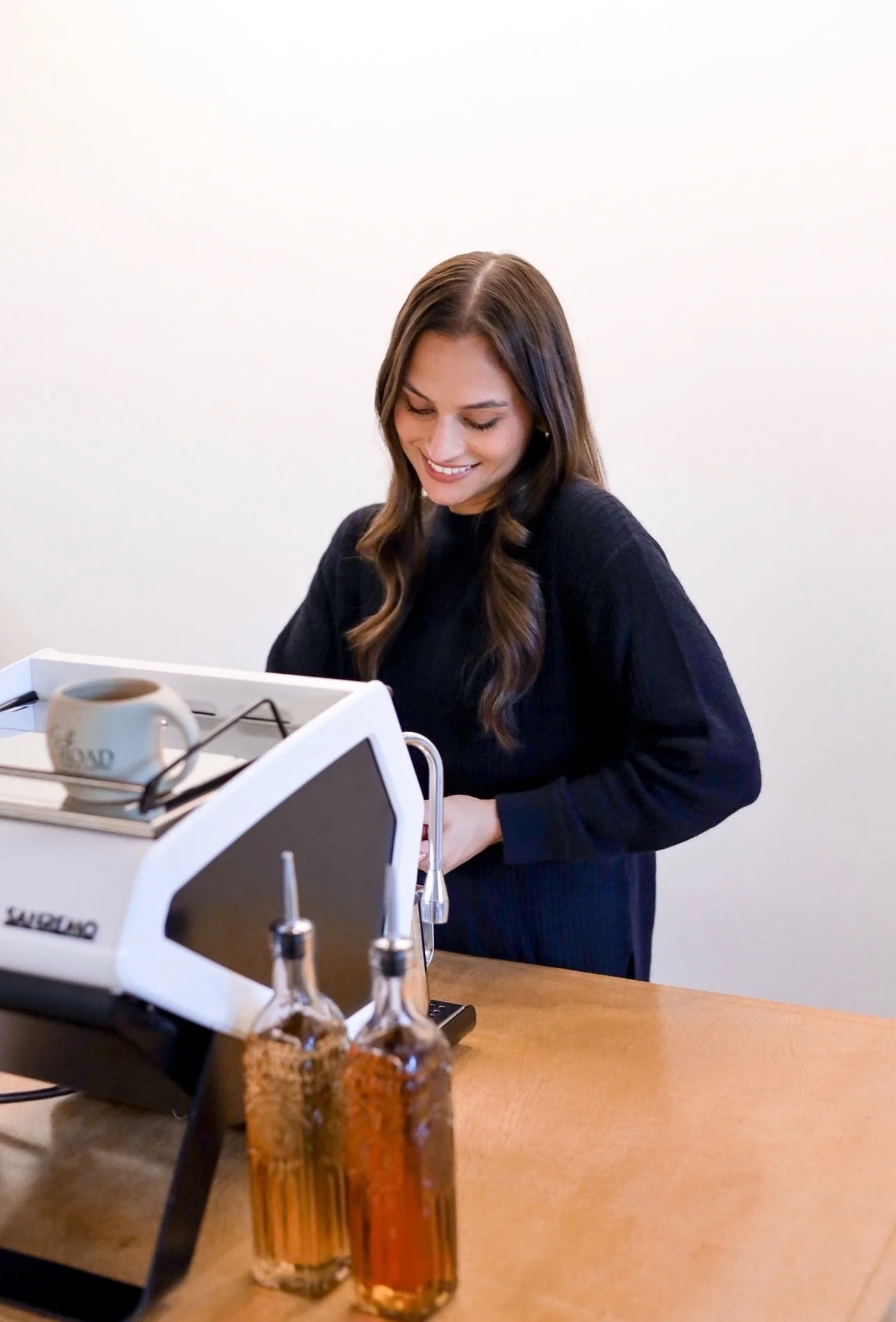 Woman with long brown hair using a machine at a wooden table, with two glass bottles of oil or vinegar and a coffee mug nearby.