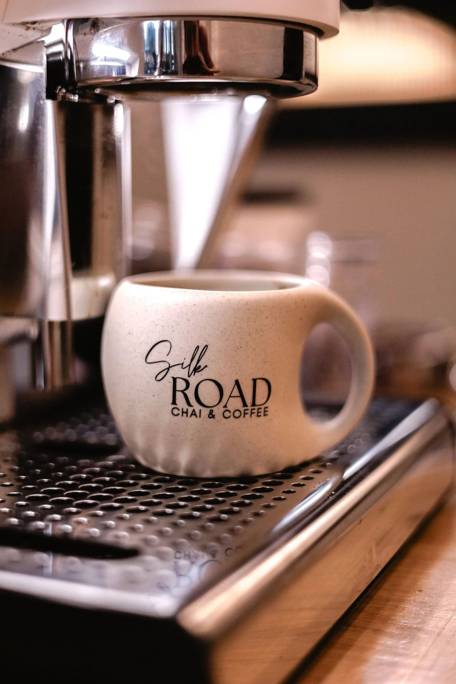 A beige coffee mug with "Silk ROAD CHAI & COFFEE" printed on it, placed under an espresso machine spout.