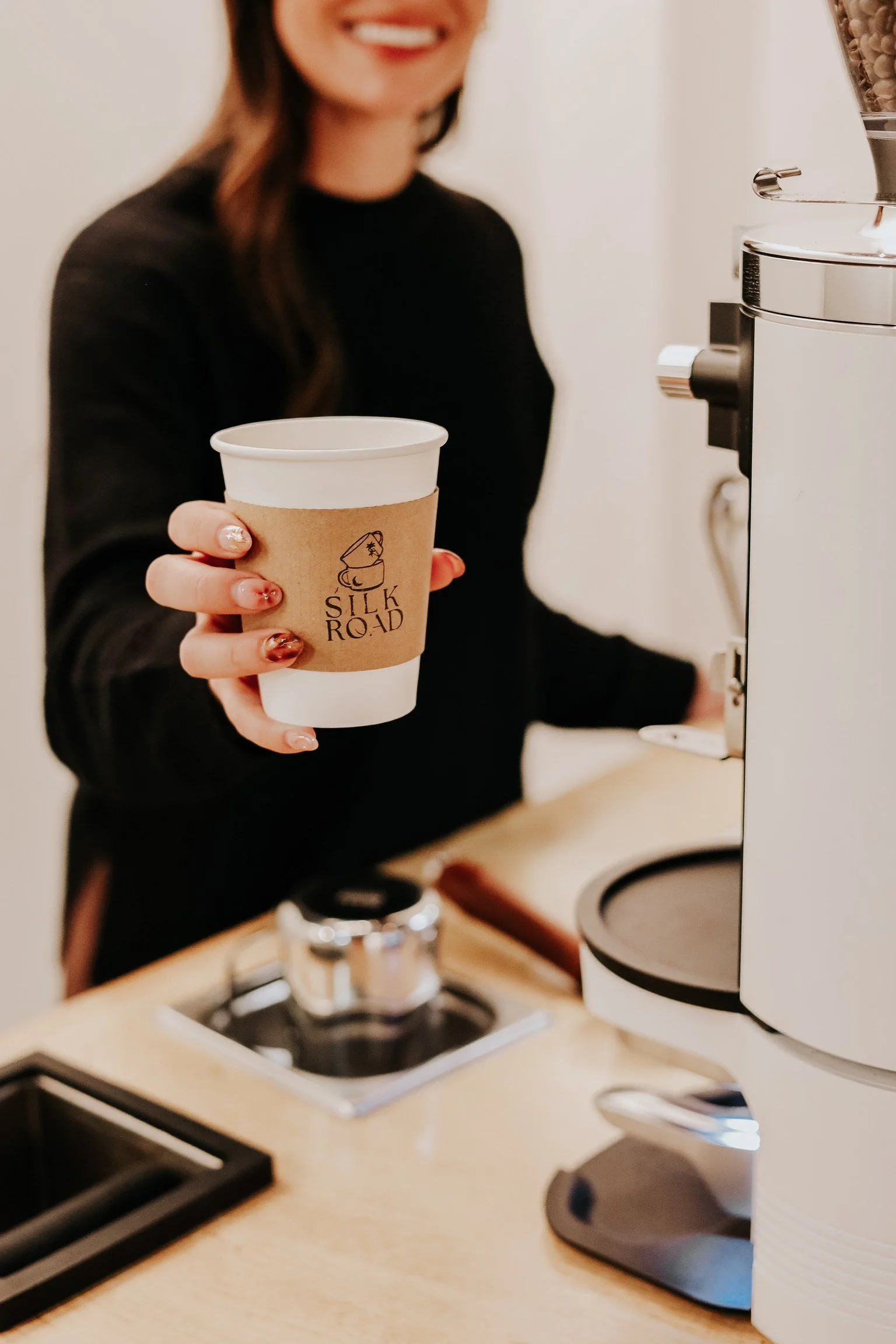 A woman holding a paper cup of coffee with a Silk Road logo, smiling, next to a coffee grinder in a cafe setting.