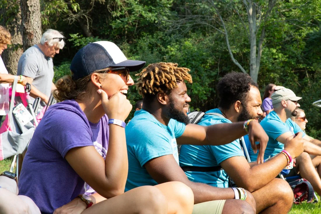 Group of diverse people sitting outdoors in a park, watching and enjoying an event, with trees and greenery in the background.