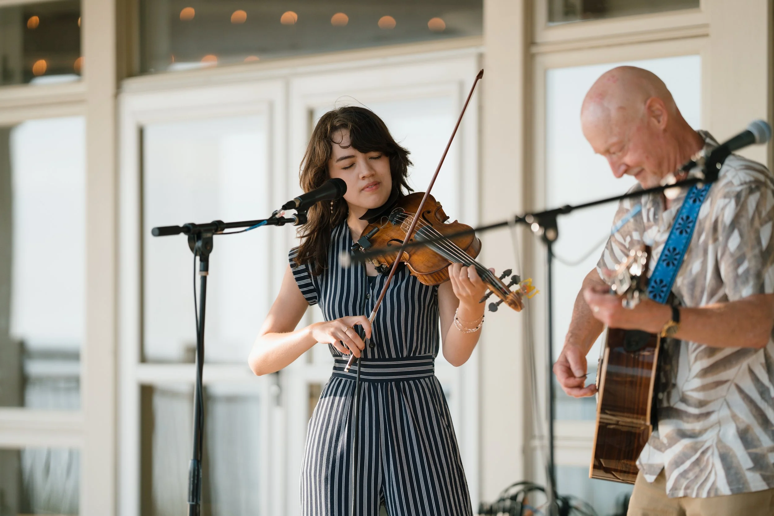 A young woman playing a violin and singing into a microphone with an older man playing guitar next to her, both performing indoors near large windows.