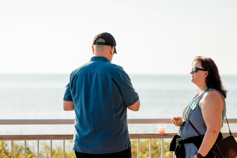 A man and a woman standing by a railing overlooking the ocean at the beach, with the man facing away from the camera and the woman facing sideways, both wearing sunglasses on a sunny day.
