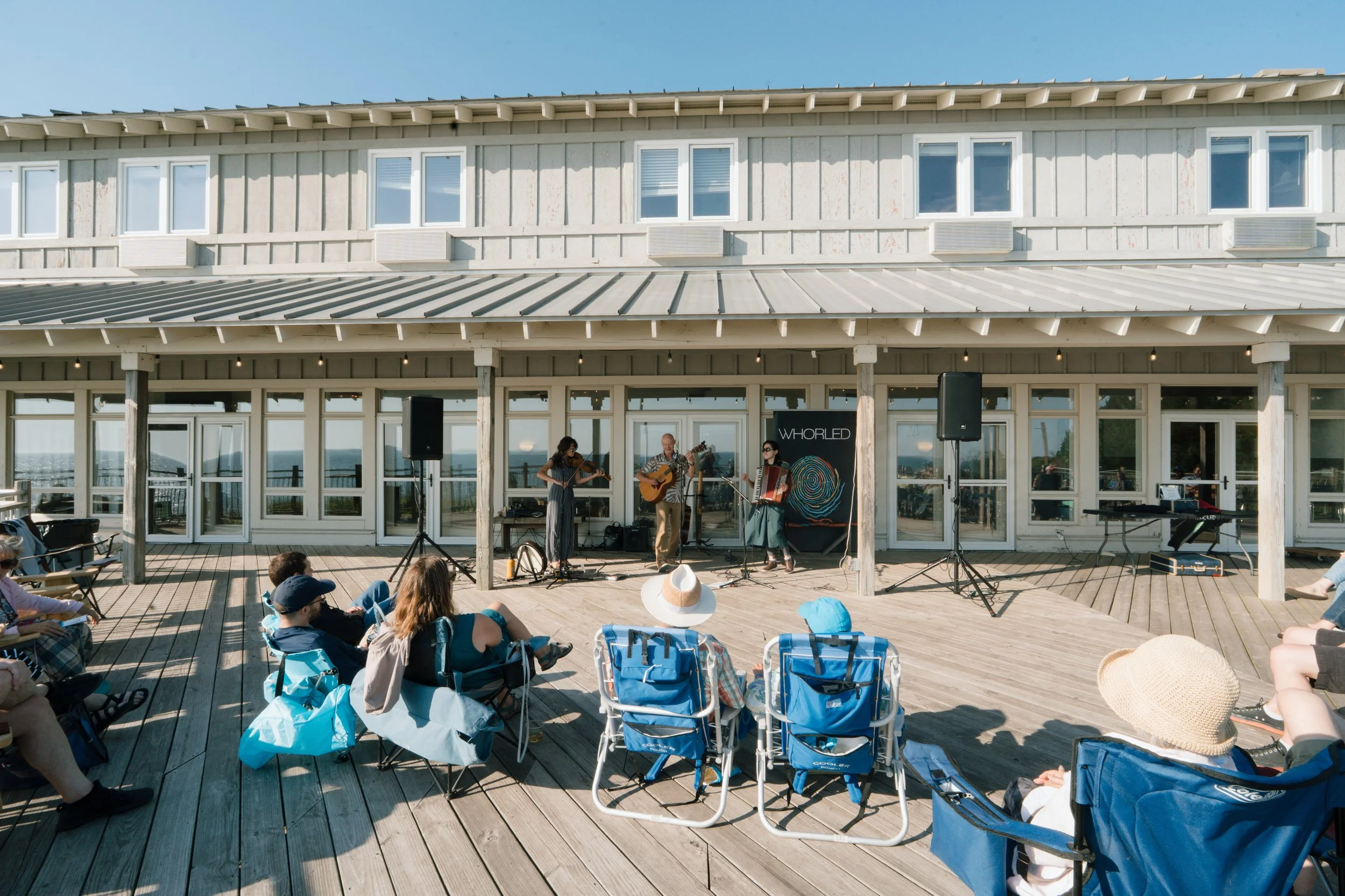 People sitting on blue chairs watching a band perform on a wooden deck outside a building with large windows and a metal roof, with the ocean in the background.