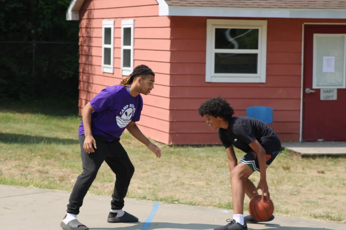 Two young men playing basketball outside on a court, with a red house in the background.