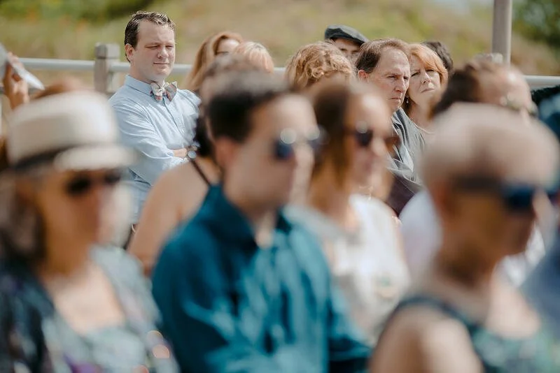 A group of people sitting outdoors, some wearing sunglasses and hats, paying attention to an event or presentation.