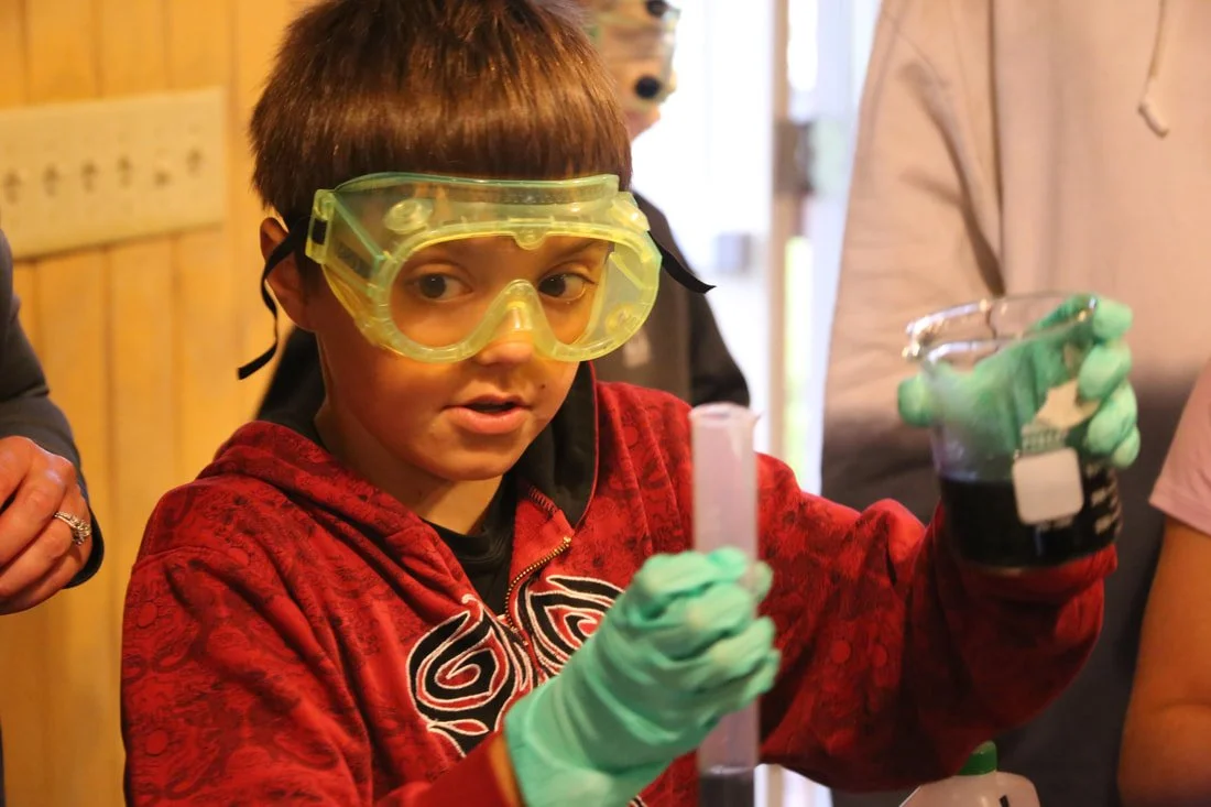 Boy wearing yellow safety goggles and green gloves, holding lab equipment in a science classroom.