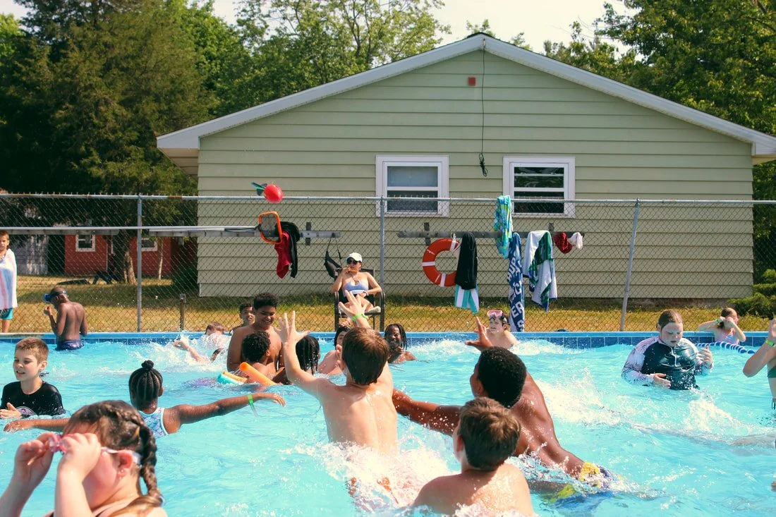 Children playing and splashing in a swimming pool during daytime, with towels, clothing hanging on a fence, and greenery in the background.