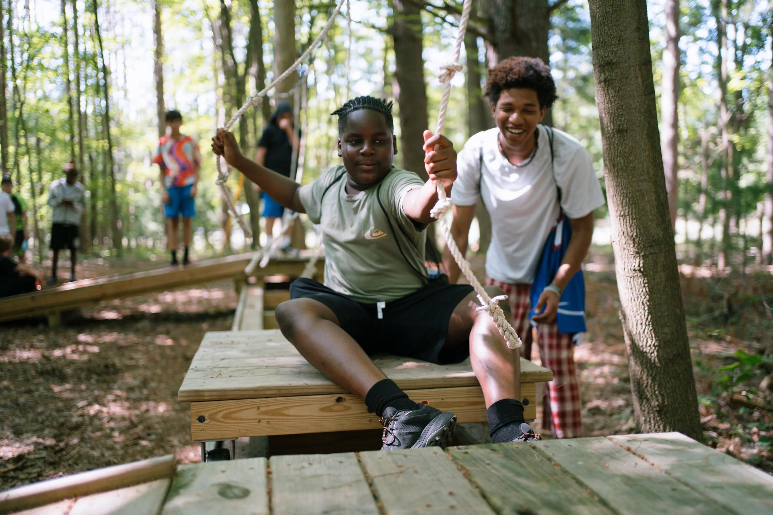 boy on a wooden flat cart pulling himself along a track with a rope. A happy counselor in the background giving support while other boys watch.