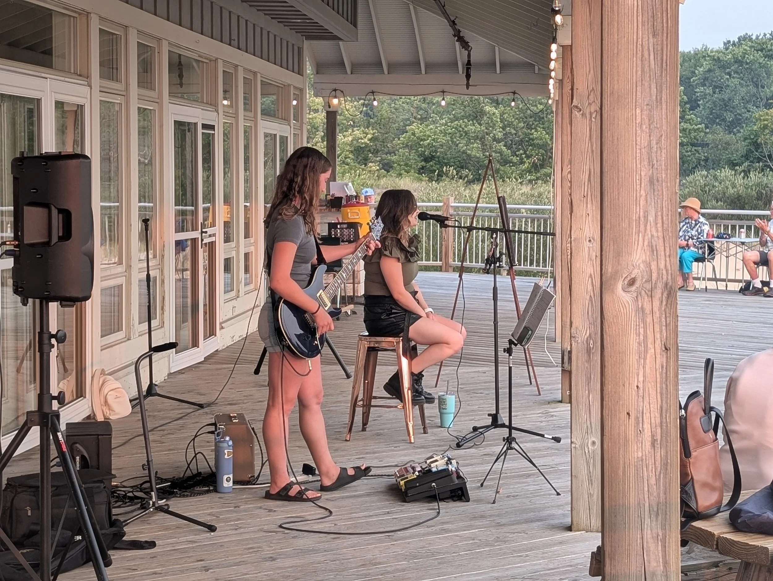 Two young women perform music outdoors on a wooden deck with a guitar and microphone, while an audience watches in the background.