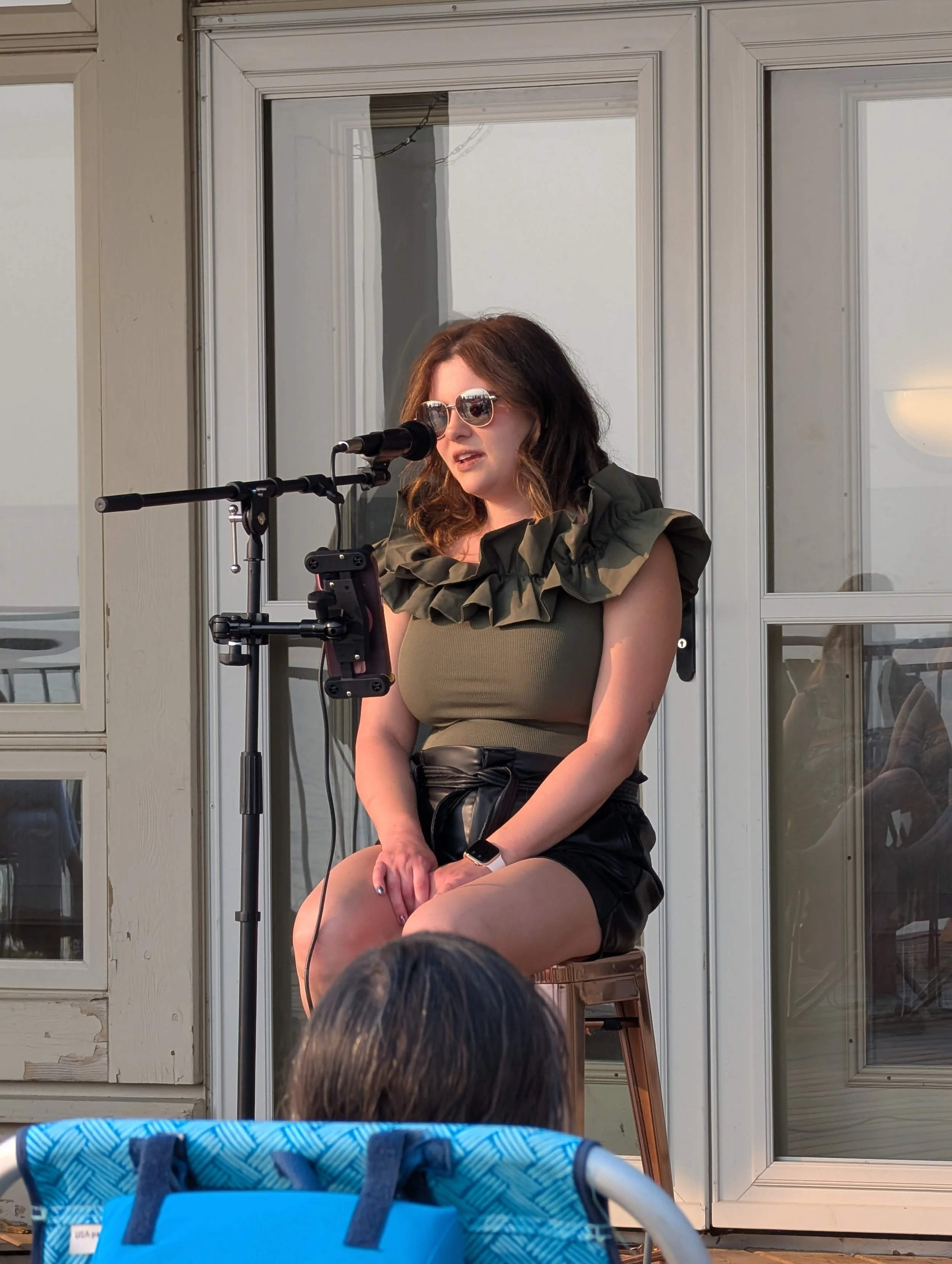 Woman sitting on a stool, speaking into a microphone, wearing sunglasses, a ruffled sleeveless top, and black shorts, with reflective windows in the background.
