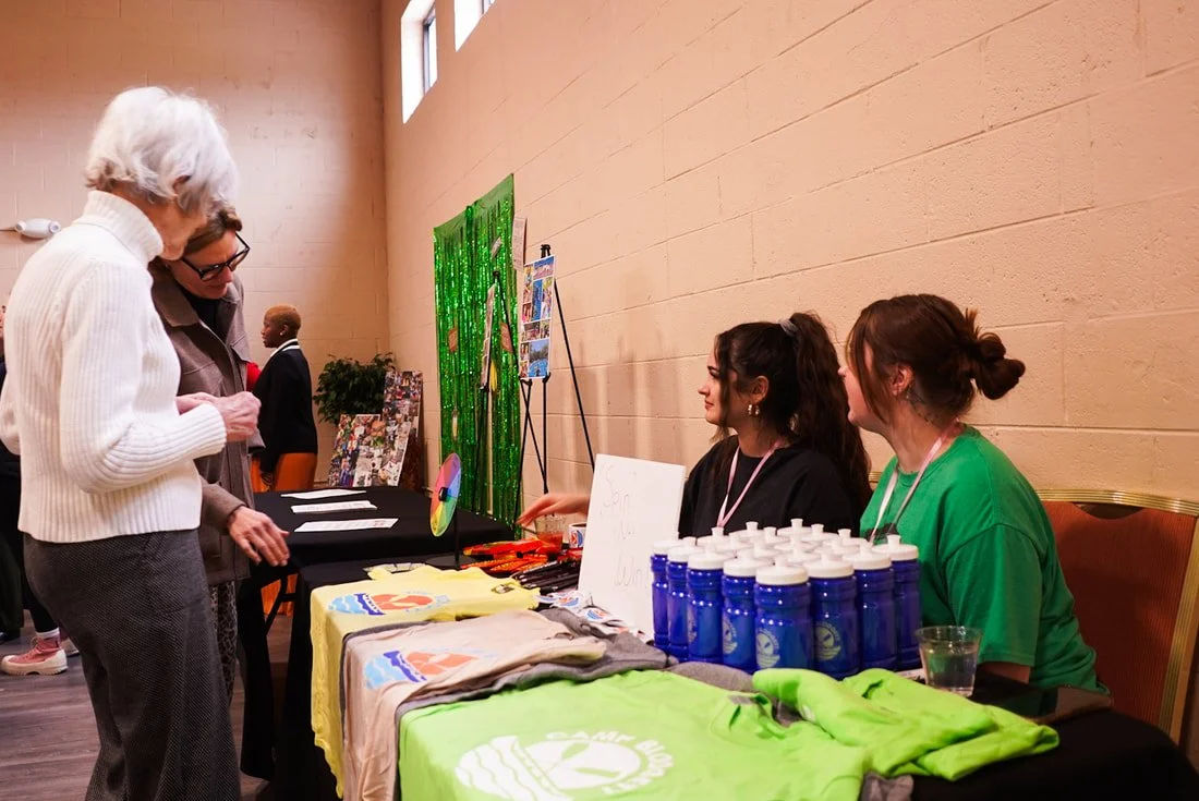 Two women sitting behind a table at an event booth, speaking to two older women standing in front of them. The table displays water bottles, T-shirts, and brochures. A green decorative backdrop and other booths are visible in the background.