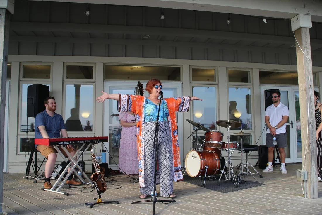 A woman with short red hair wearing sunglasses, a blue top, and an orange, blue, and white patterned cover-up, stands on a wooden stage with her arms outstretched, singing into a microphone. She is surrounded by four band members, including a keyboar