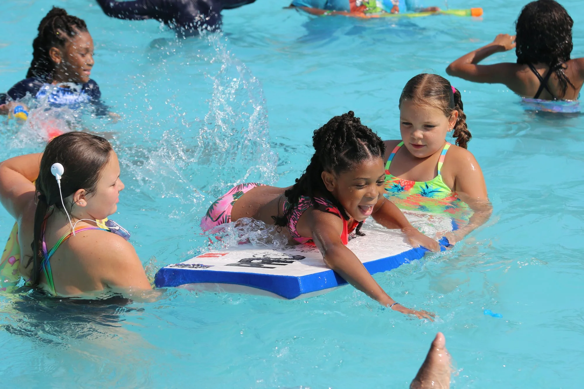 Children enjoying swimming lesson at the pool, with one girl on a kickboard and others splashing and playing in the water.