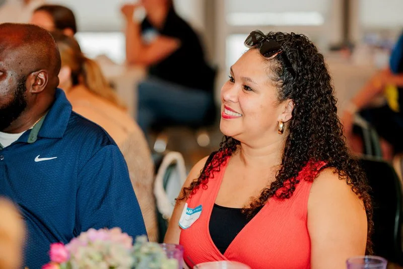 A woman with curly hair, wearing sunglasses on her head and a sleeveless red top, smiling and sitting at a gathering with other people in the background.
