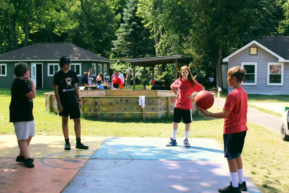 Kids playing basketball outdoors, with a group of children in the background near a small building and trees.