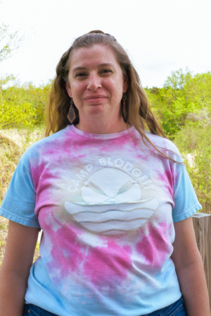A woman standing outdoors in front of green trees, wearing a multicolored tie-dye t-shirt with a boat and water design on it.