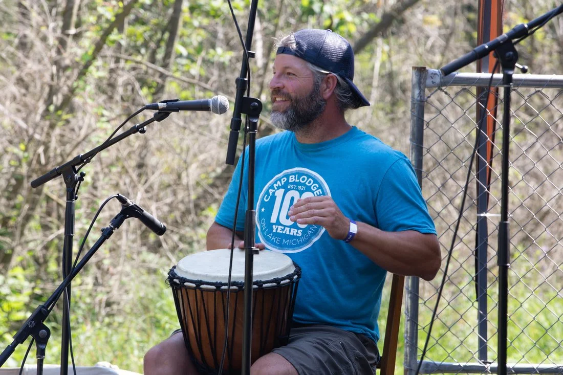 A man with a beard and wearing a blue Camp Blodgett T-shirt and a backwards baseball cap, smiling while playing a djembe drum outdoors near a microphone.