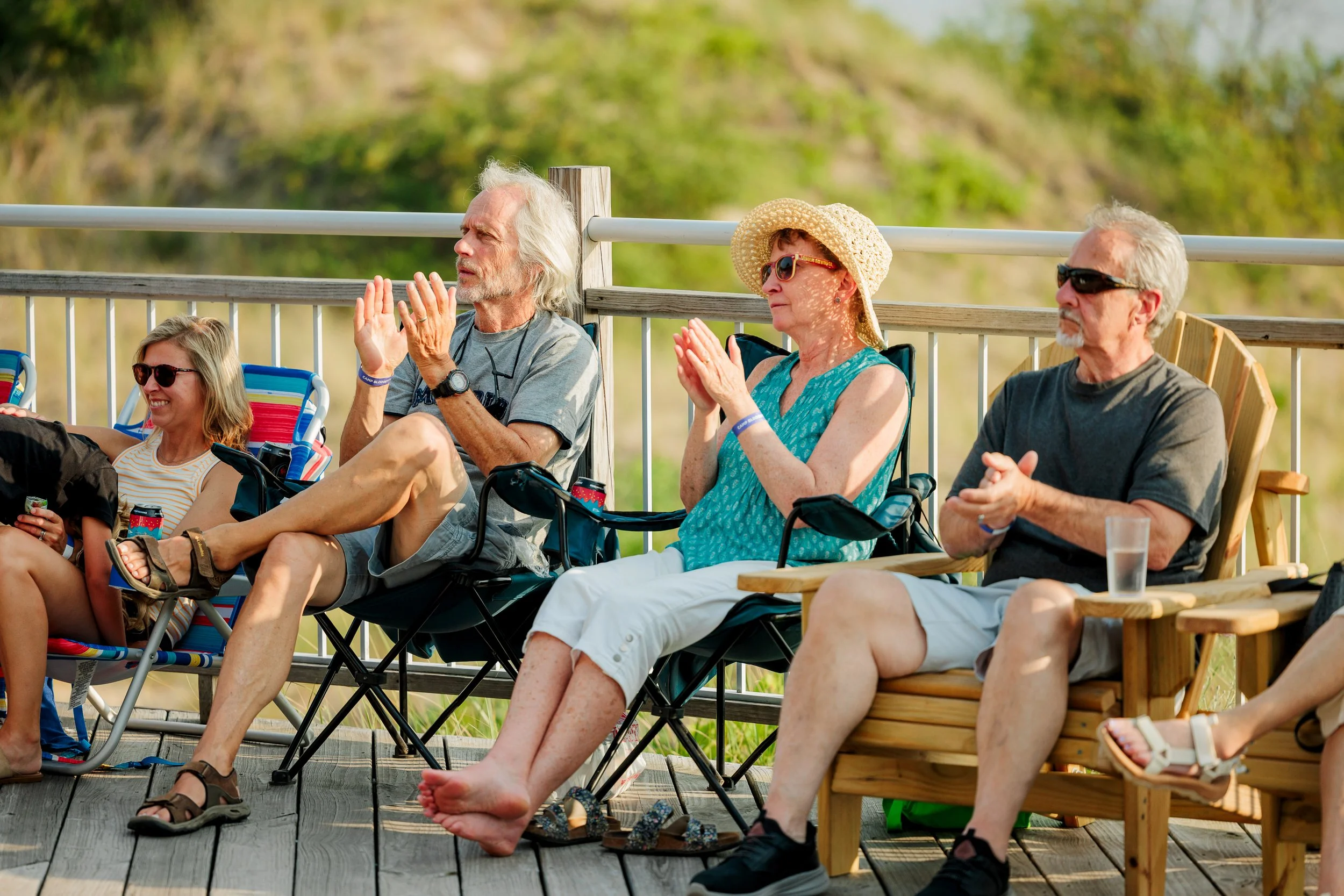 People sitting outdoors on a deck, clapping and enjoying a sunny day, with greenery in the background.