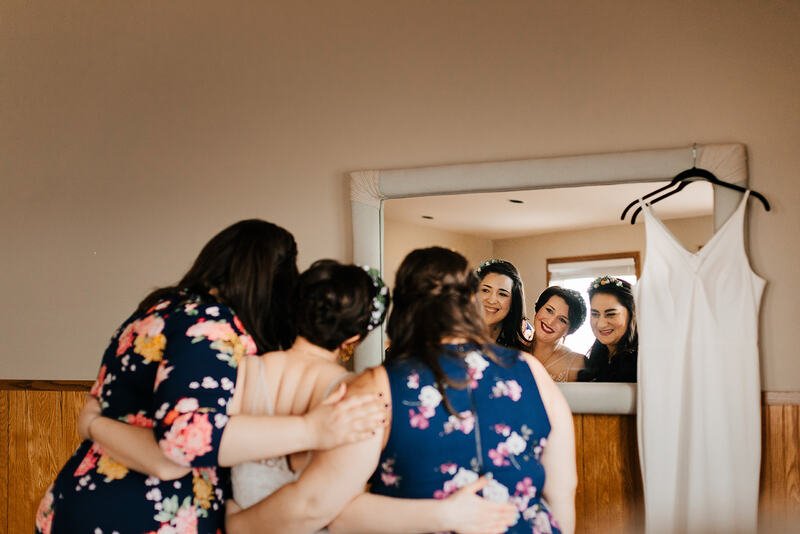 Four women looking into a mirror, smiling, with a wedding dress hanging next to it.