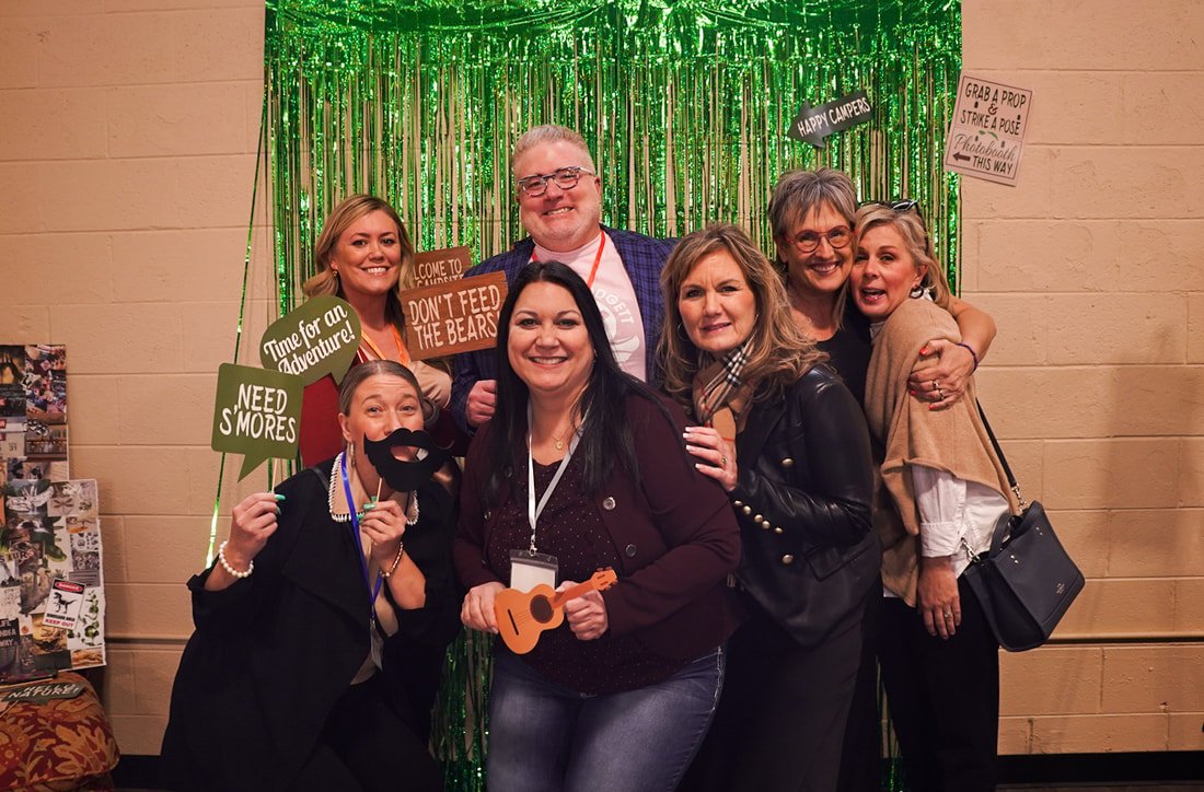 Six women and one man smiling at a photo booth with green shiny fringe backdrop, some holding props such as signs and a puppet mustache, with signs reading 'Time for an Adventure!', 'NEED S'MORES', and 'DON'T FEED THE BEARS'.