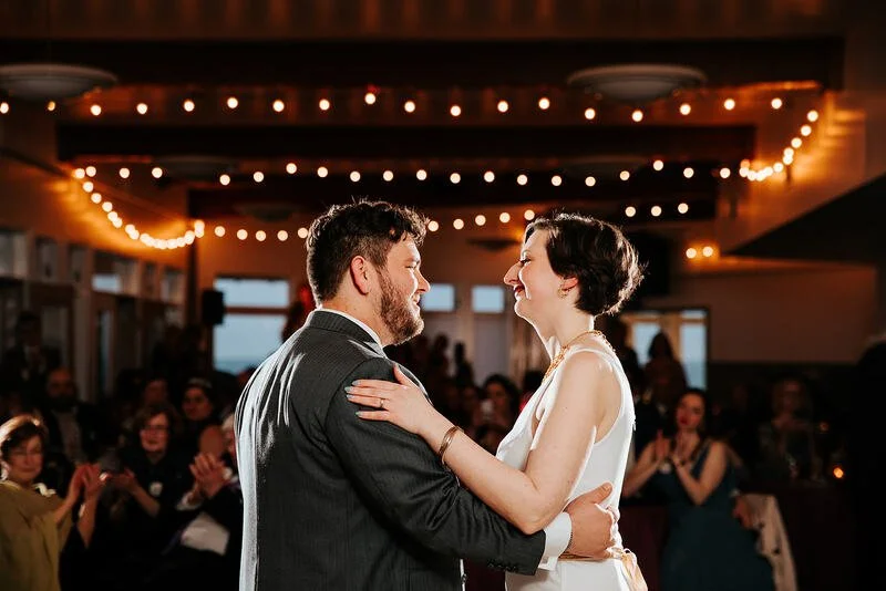 A couple is dancing and smiling at their wedding reception with wedding guests clapping in the background, decorated with string lights.