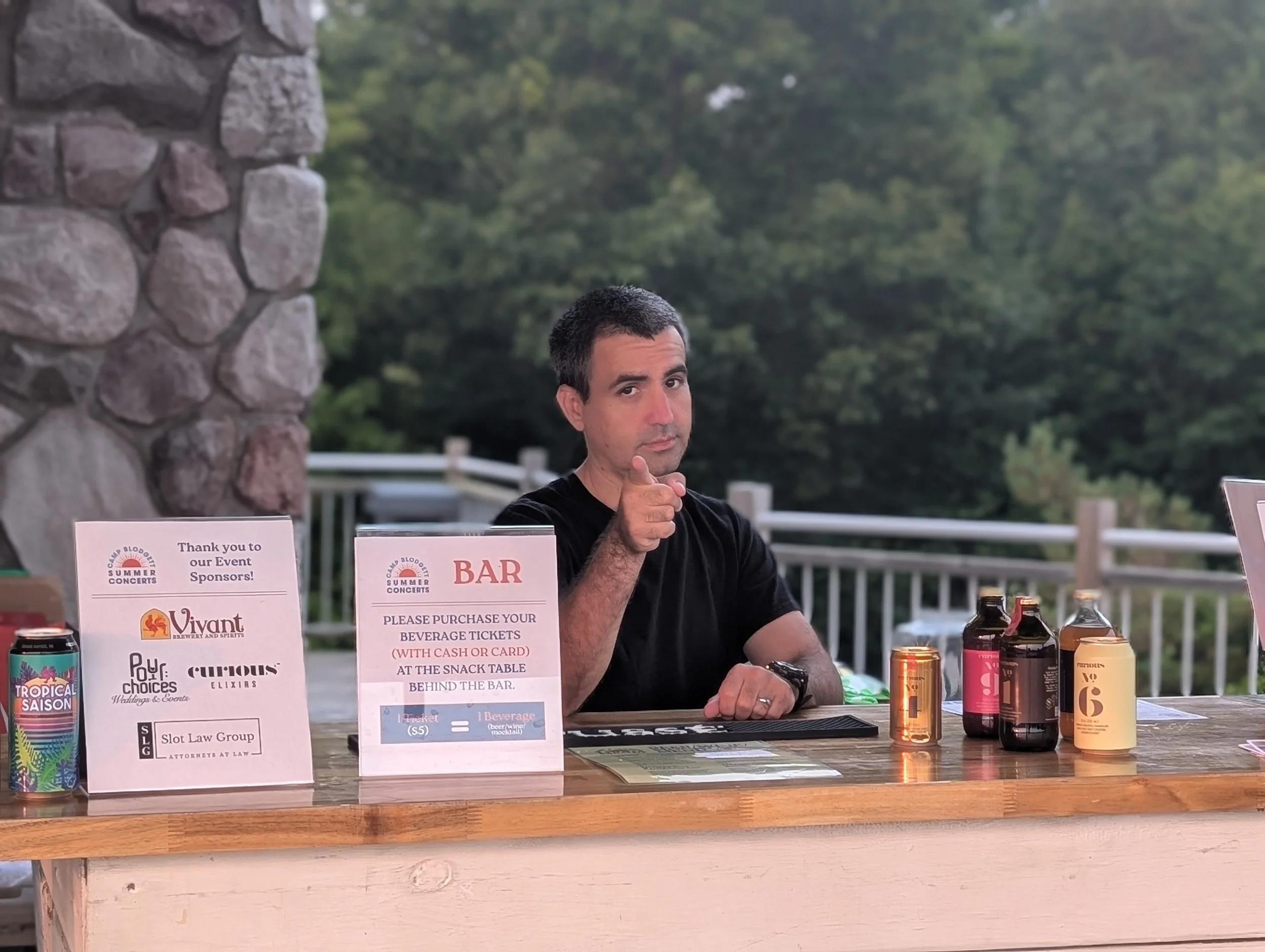 A man sitting at a bar counter outdoors, pointing towards the camera with a playful expression. Various drink bottles and cans are on the counter, along with signs and a laptop. Trees and a fence are visible in the background.
