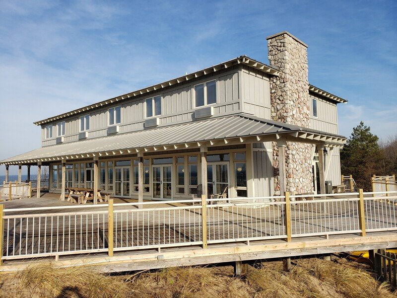 A two-story house with a gray exterior, large glass windows, and a stone chimney, featuring a wraparound deck with outdoor seating and a picnic table, set in a natural environment under a blue sky.