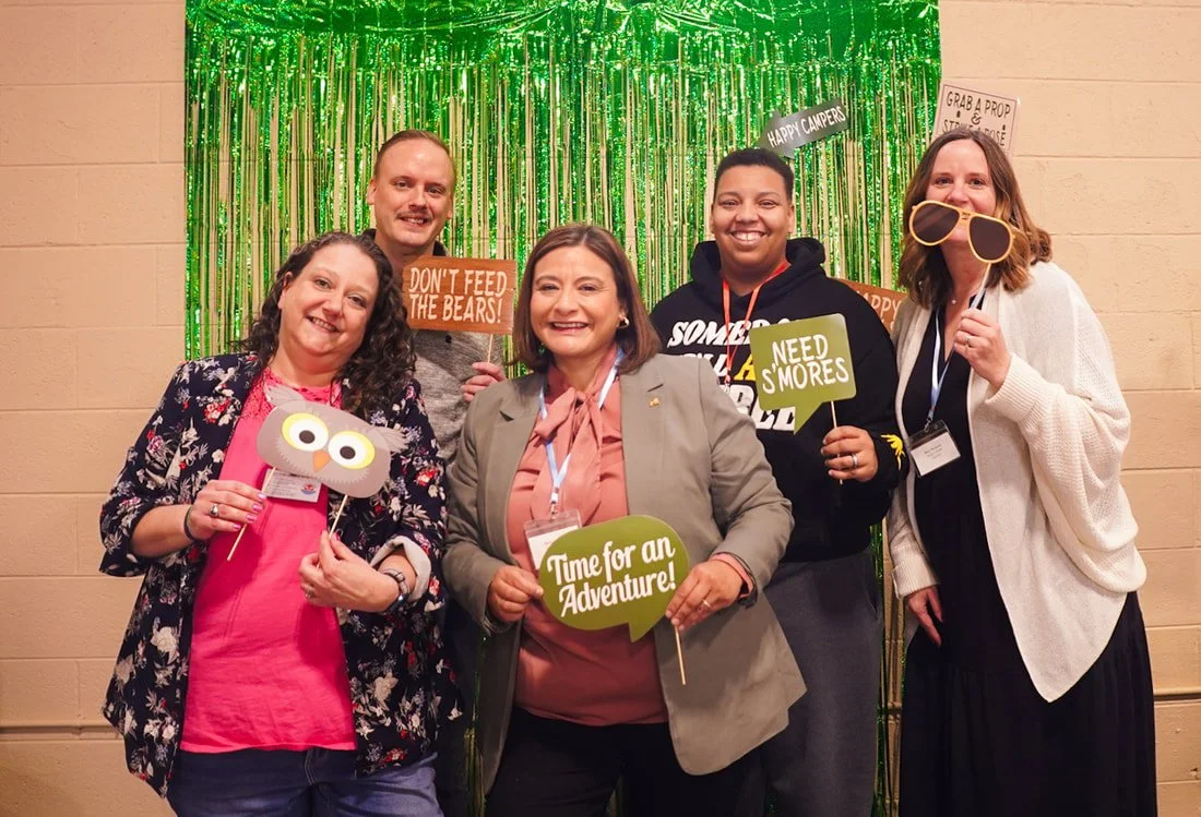 Five people smiling at a photo booth with a green metallic fringe backdrop, holding props like glasses, speech bubbles, and signs with messages such as "Don't Feed the Bears!", "Need S'mores", and "Time for an Adventure!"