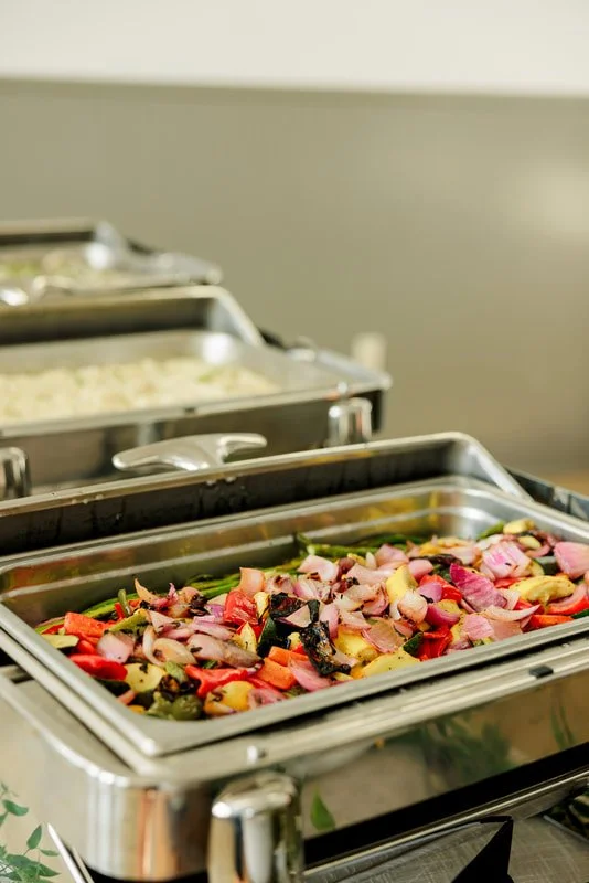 A stainless steel chafing dish filled with roasted vegetables, including red bell peppers, onions, and zucchini, in a buffet setting.