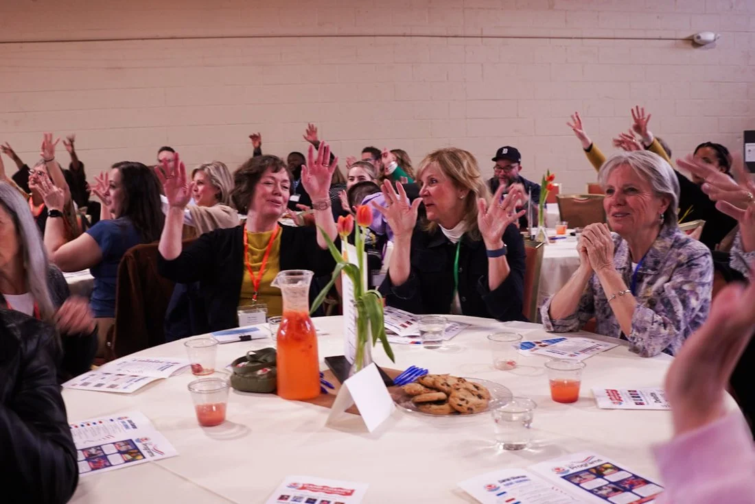 People seated around a table raising their hands, enjoying a social event with cookies and drinks, with a flower centerpiece.