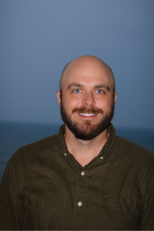A smiling man with a beard and bald head, wearing a dark green button-up shirt, standing outdoors with a cloudy sky and ocean in the background.
