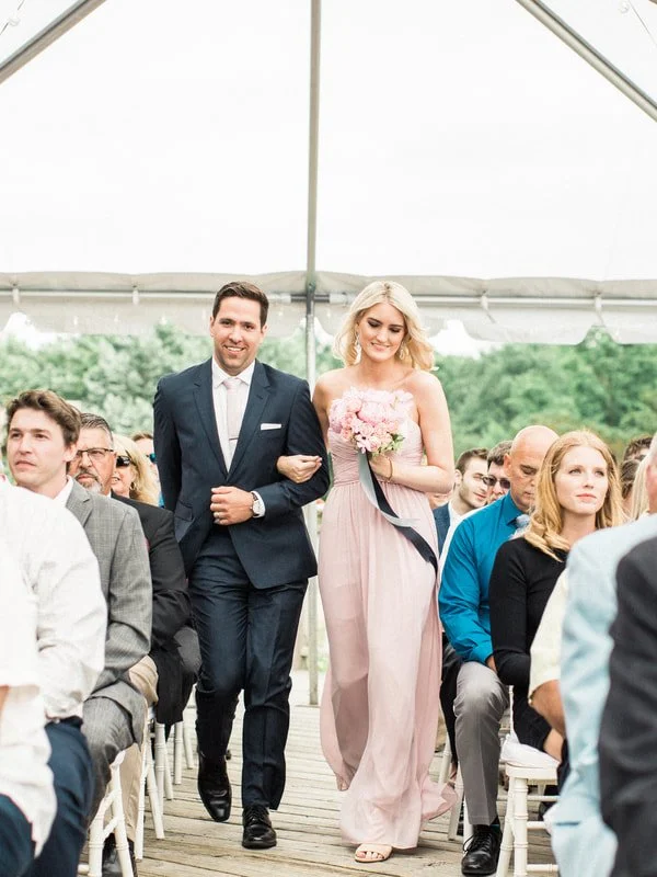 A couple walking down the aisle at an outdoor wedding ceremony, with guests seated on either side under a canopy.